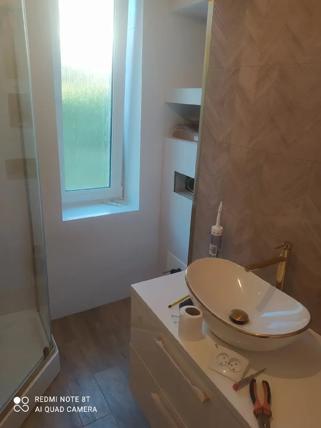 Nowoczesna łazienka w trakcie wykańczania: owalna umywalka nablatowa z złotymi akcentami, płytki imitujące drewno, białe meble i okno. Widoczne narzędzia.