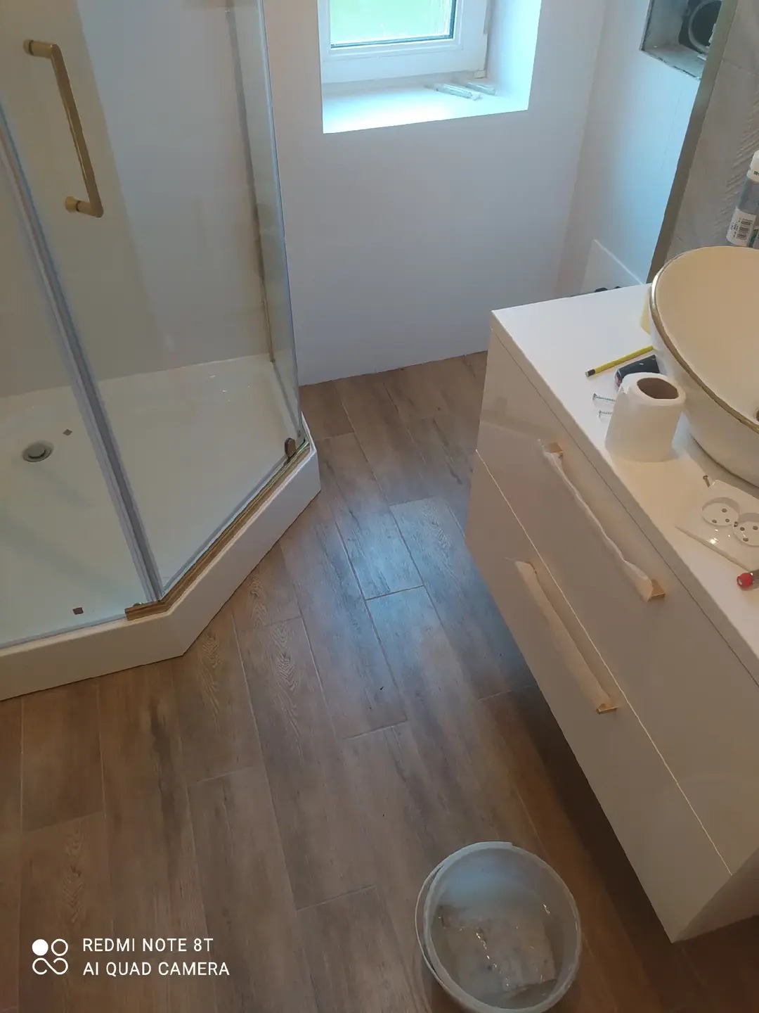 Wykończona łazienka z kabiną prysznicową z złotymi detalami, białą szafką z umywalką i drewnopodobną podłogą. Widoczne narzędzia i akcesoria.