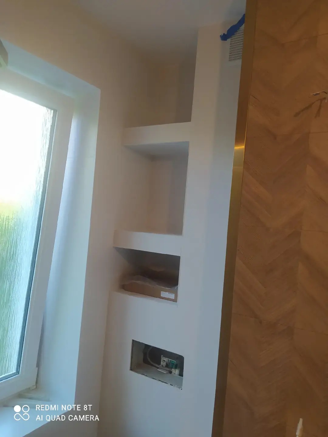 Biała ściana z wnękami w łazience po remoncie, widoczne instalacje i okno. Na ścianie dekoracyjna okładzina imitująca drewno ułożona w jodełkę.