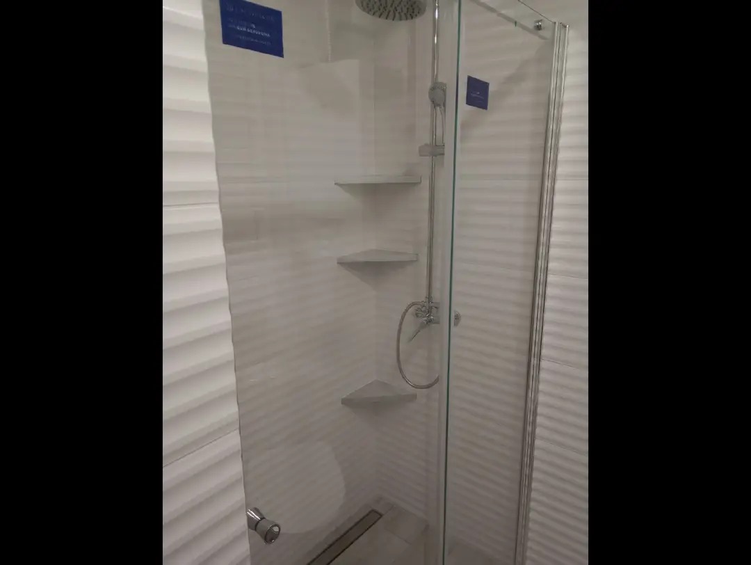 Nowoczesna kabina prysznicowa z półkami narożnymi i deszczownicą, wykończona białymi płytkami z delikatną fakturą fal. Minimalistyczny design.