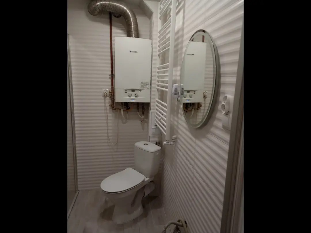 Wyremontowana łazienka w Kętrzynie z toaletą, grzejnikiem drabinkowym i piecem gazowym. Ściany wykończone w jasnej kolorystyce z poziomymi paskami.