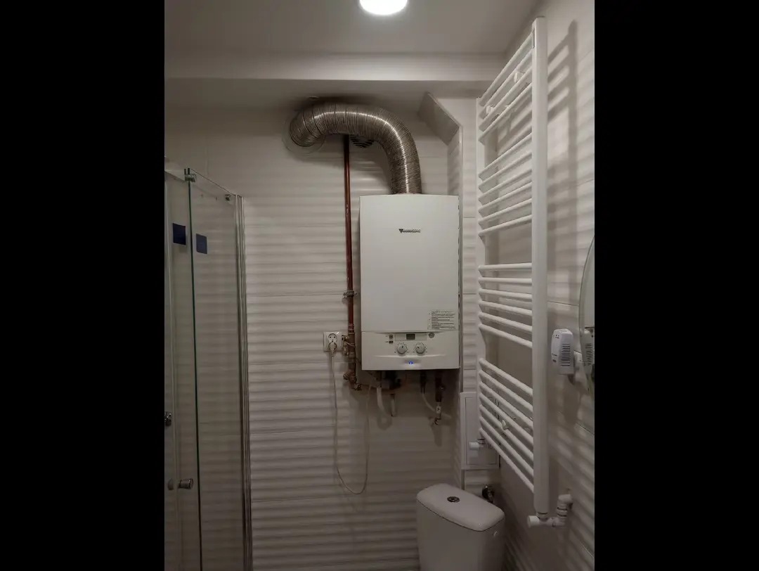 Biały piec gazowy z rurą odprowadzającą spaliny w łazience z grzejnikiem drabinkowym i kabiną prysznicową. Widoczna toaleta i płytki imitujące drewno na ścianie.