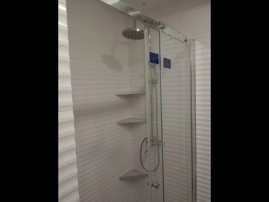 Nowoczesna kabina prysznicowa z deszczownicą i półkami narożnymi, wykończona białymi płytkami z subtelną fakturą. Minimalistyczny design, chromowane detale.