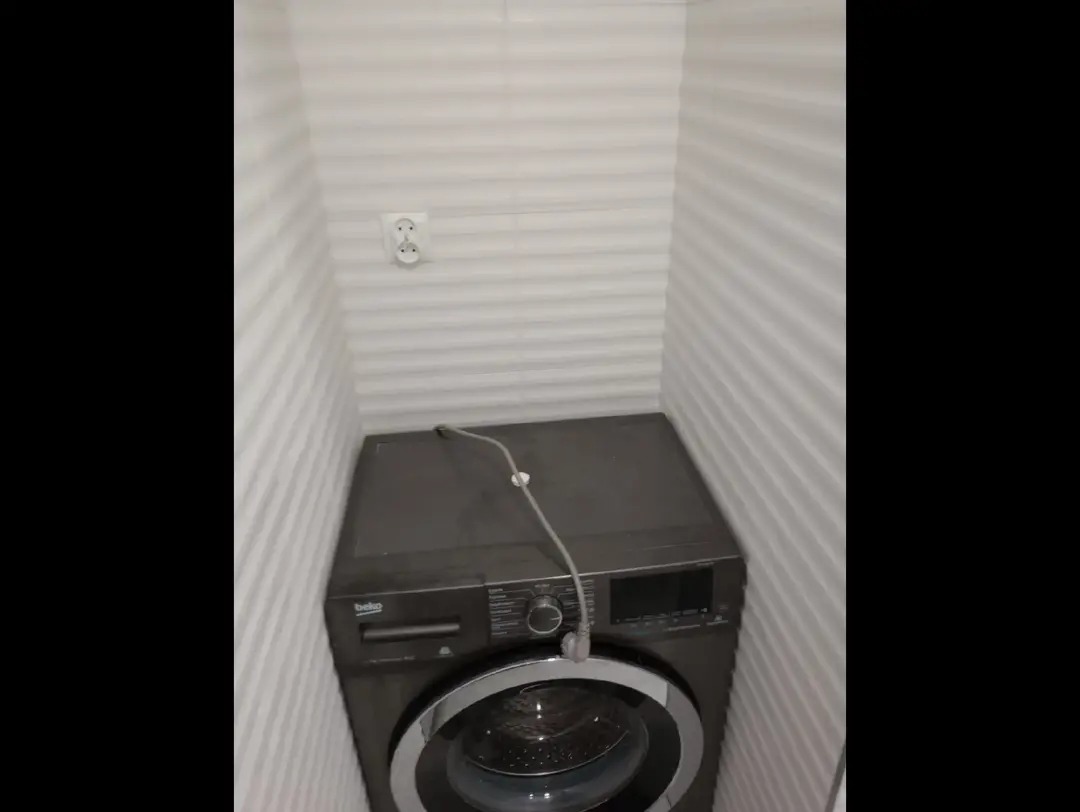 Ciemna pralka Beko w małej, wykończonej białymi panelami łazience. Widoczne gniazdko elektryczne i kabel zasilający. Ujęcie z góry, ciasna przestrzeń.