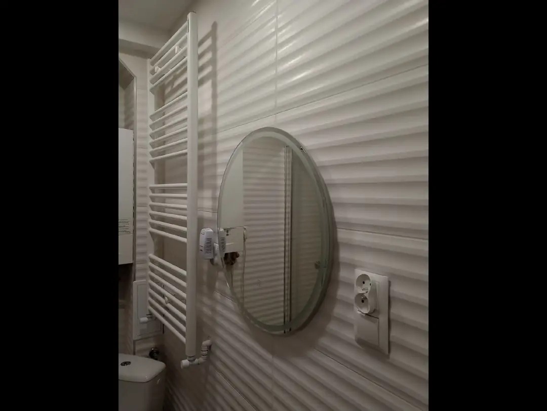 Biała łazienka z grzejnikiem drabinkowym, owalnym lustrem i falistymi płytkami. Widoczny fragment toalety i gniazdko elektryczne. Nowoczesny design.