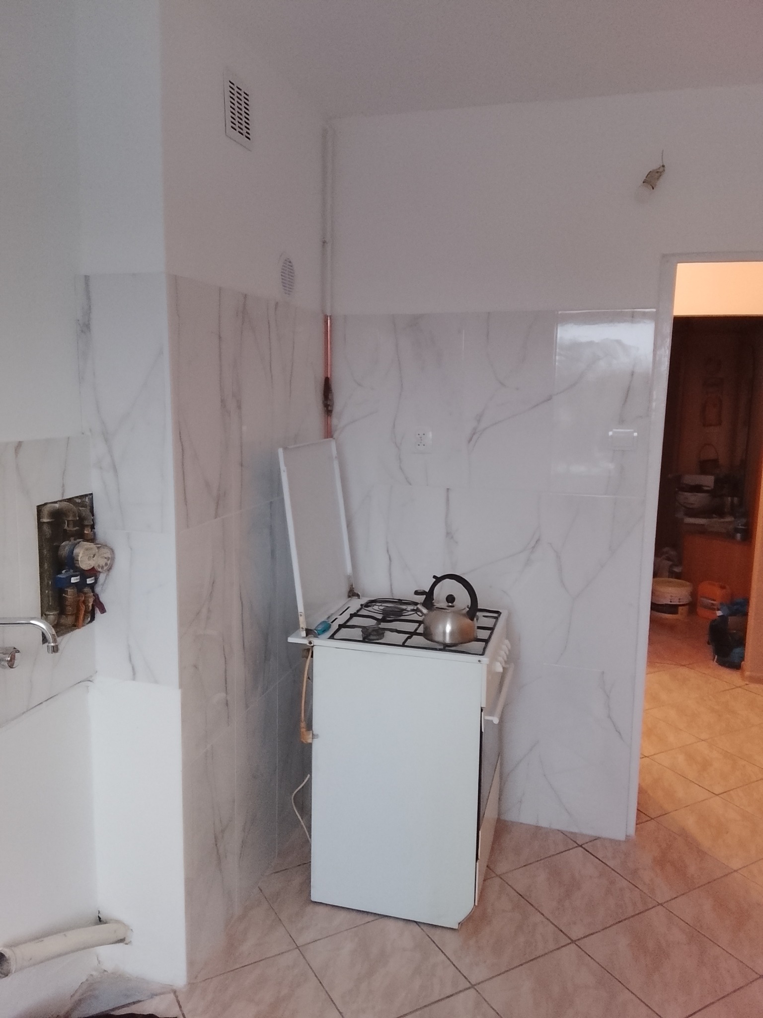 Wykończona kuchnia z białymi płytkami imitującymi marmur. Widoczna kuchenka gazowa z czajnikiem, fragment instalacji wodnej i wejście do innego pomieszczenia.