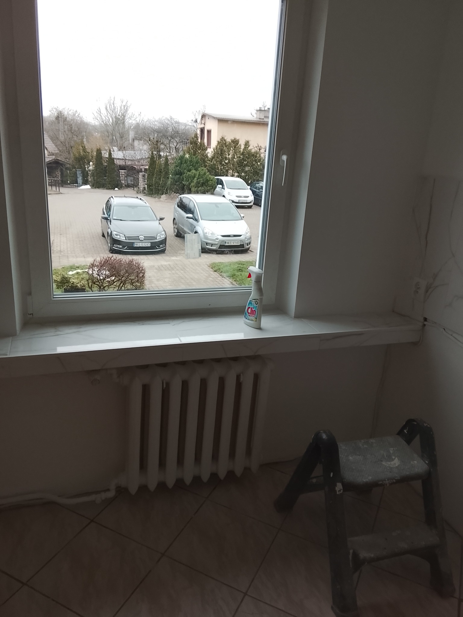 Wnętrze pokoju z oknem, widok na podjazd z samochodami. Na parapecie płyn do czyszczenia, pod oknem grzejnik i drabinka malarska. Widoczne ślady prac wykończeniowych.