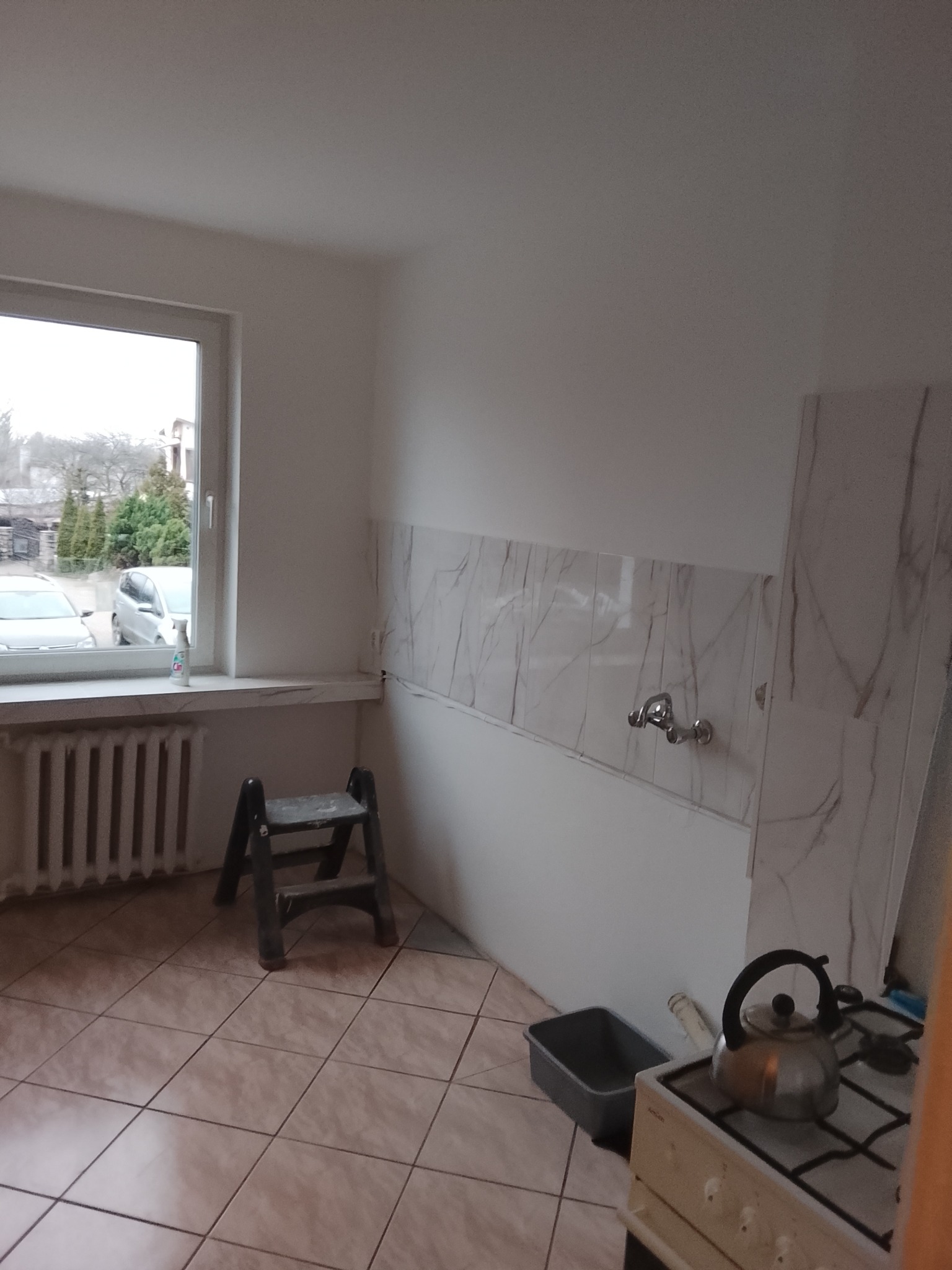 Fragment kuchni w trakcie remontu: częściowo położone marmurowe płytki nad zlewem, drabina, grzejnik i kuchenka z czajnikiem. Widok z okna na zewnątrz.