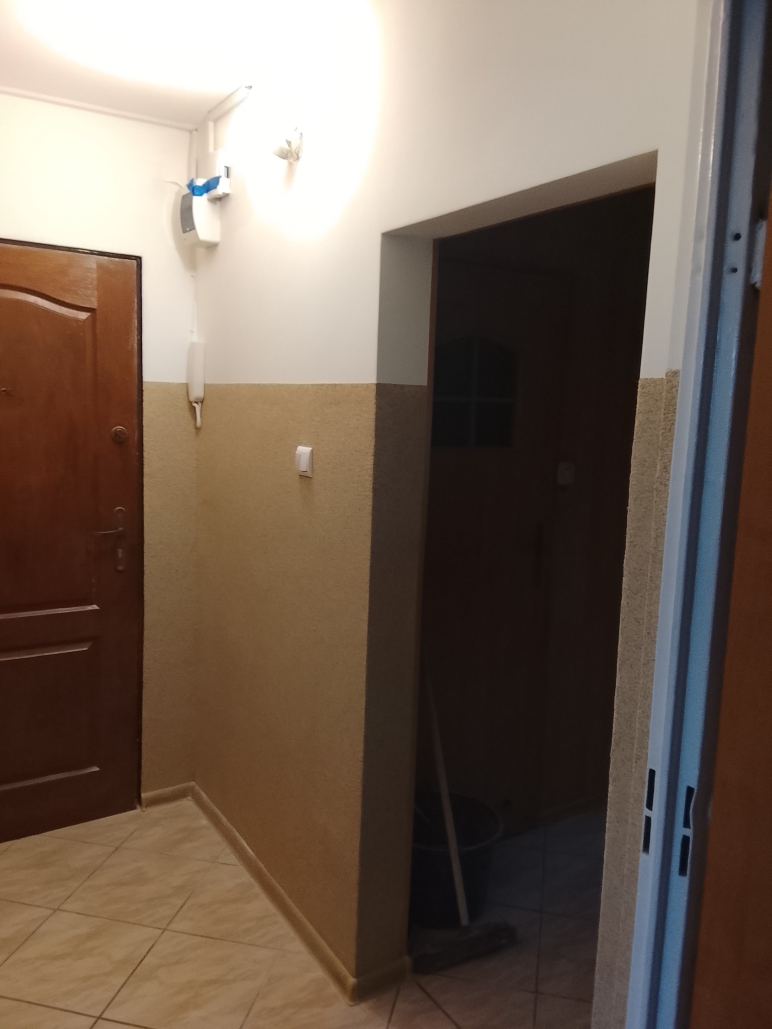 Korytarz po odświeżeniu: pomalowane ściany w dwóch odcieniach beżu, brązowe drzwi wejściowe, płytki podłogowe w ciepłych tonach. Widoczny fragment otwartych drzwi wewnętrznych.