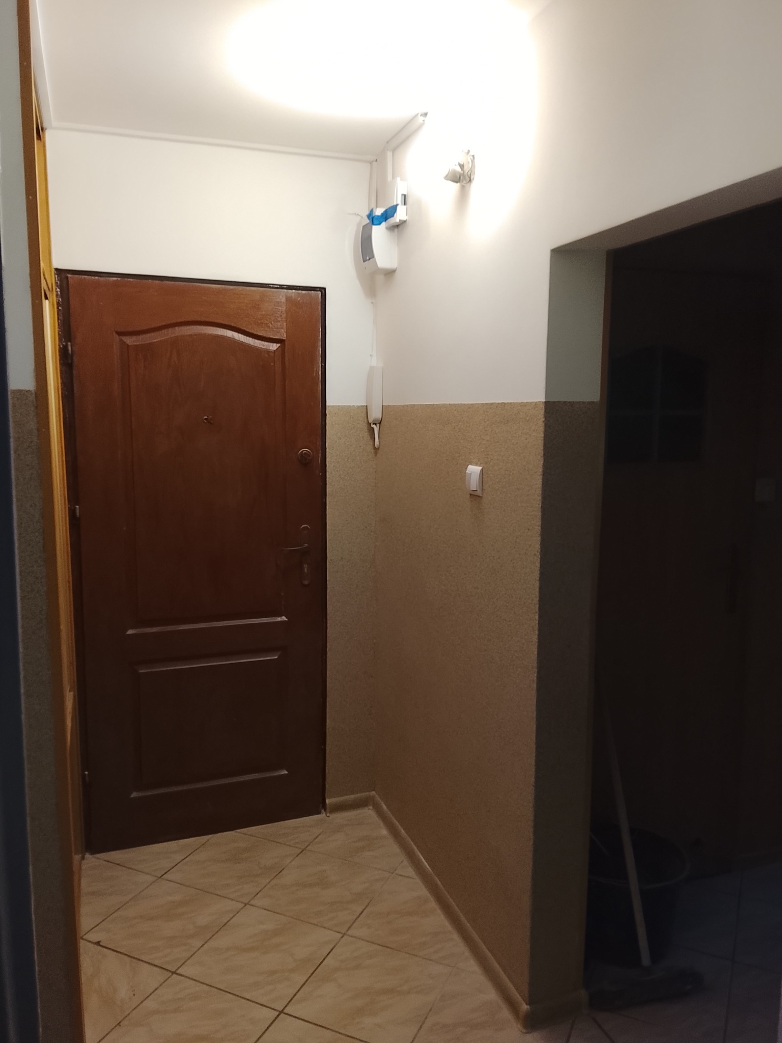 Ciemne, drewniane drzwi wejściowe w korytarzu z beżowymi płytkami podłogowymi i ścianami w dwóch odcieniach. Widoczny domofon i włącznik światła.