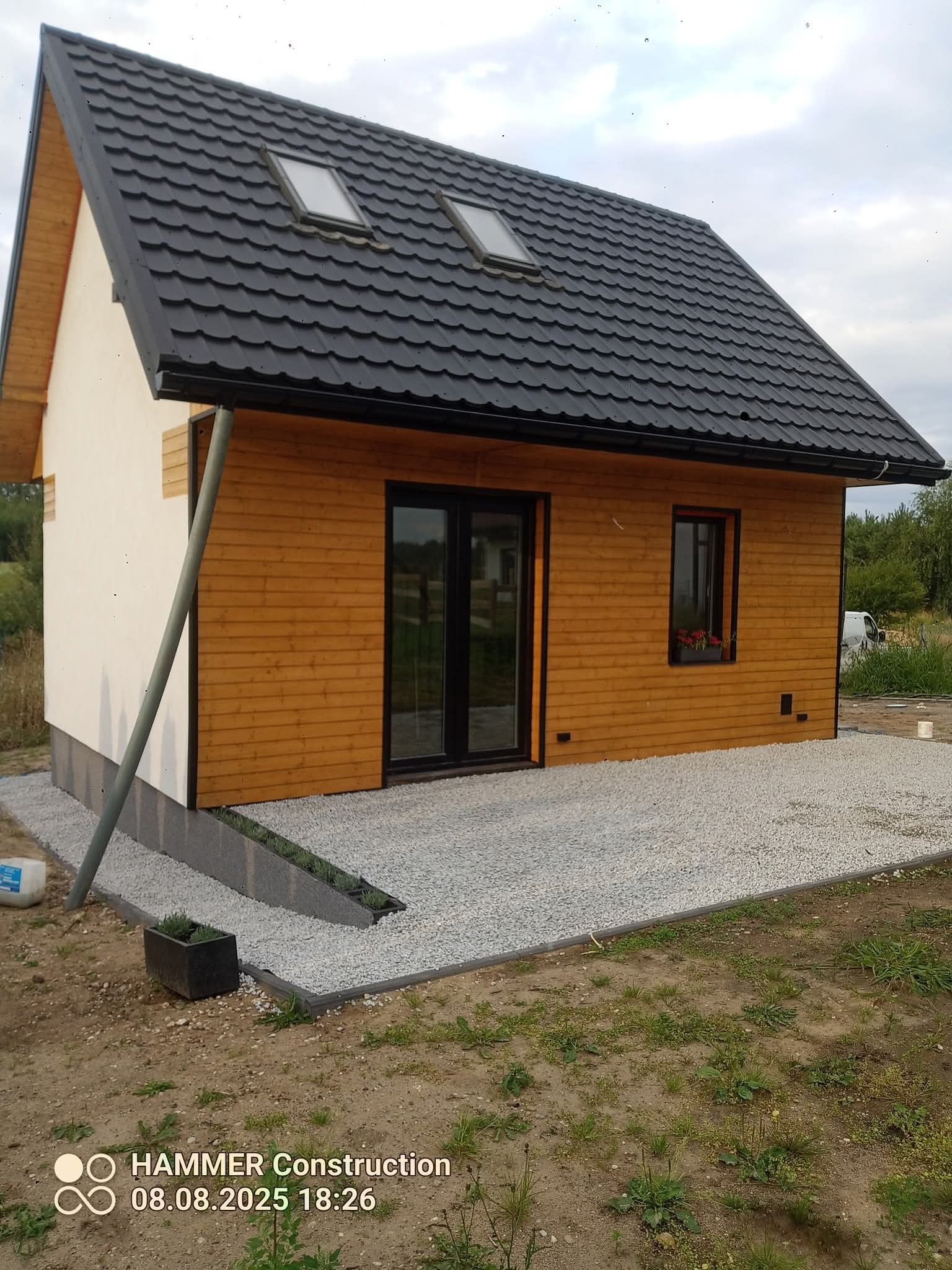 Nowoczesny dom z drewnianą elewacją i czarnym dachem, widok z zewnątrz. Uwagę przyciągają okna dachowe i kamienny podjazd. Minimalistyczny styl.