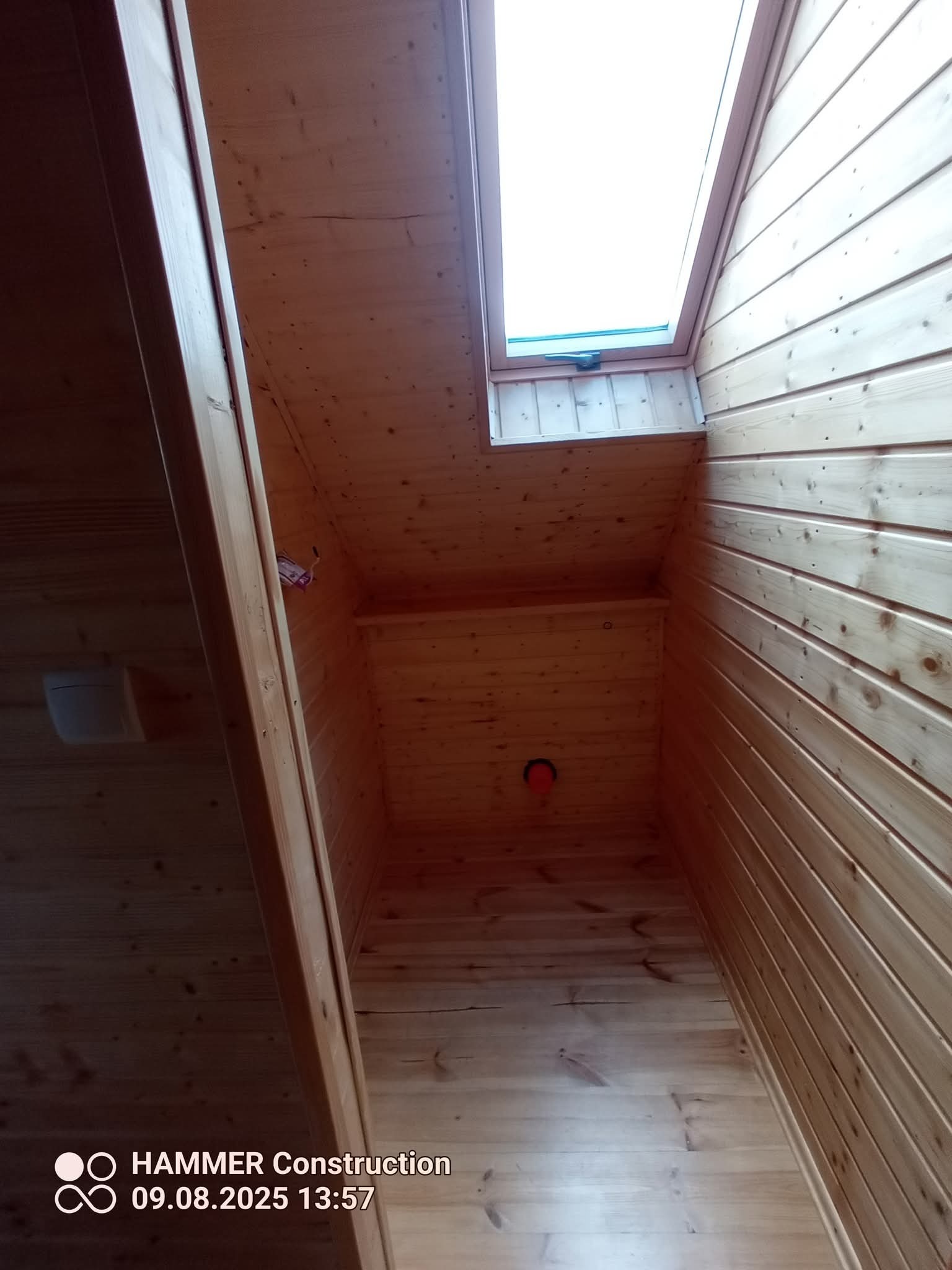 Drewniane panele boazeryjne na poddaszu z oknem dachowym. Widoczne łączenia paneli i otwór instalacyjny. Ujęcie z dołu, podkreślające geometrię wnętrza.