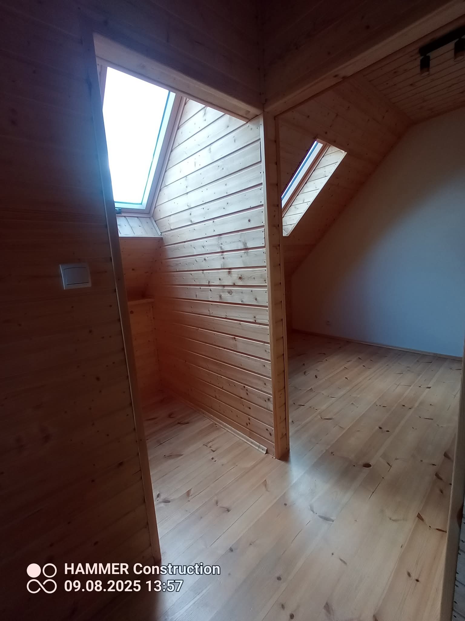 Drewniane wykończenie poddasza z oknem dachowym i podłogą z desek. Widok z korytarza na jasne, wykończone drewnem wnętrze.