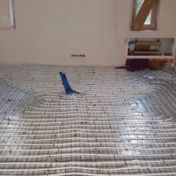 MK Hydro Instal Bud - Białe rury ogrzewania podłogowego ułożone na folii aluminiowej, z niebieskim elementem montażowym i rozdzielaczem w ścianie, w surowym wnętrzu.