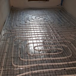 MK Hydro Instal Bud - Ułożone rury ogrzewania podłogowego na folii z siatką, widoczne mocowania. W tle okno i fragment instalacji na ścianie. Etap przed wylaniem posadzki.