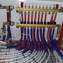 MK Hydro Instal Bud - Rozdzielacz ogrzewania podłogowego z rurami w kolorach czerwonym i niebieskim, zamontowany w białej szafce, widoczne białe rury na podłodze z siatką.