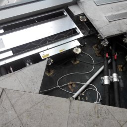 MK Hydro Instal Bud - Otwarta podłoga techniczna z widoczną instalacją: rury, kable i metalowa skrzynka. Fragment buta w rogu kadru. Widok z góry na instalację pod podłogą.