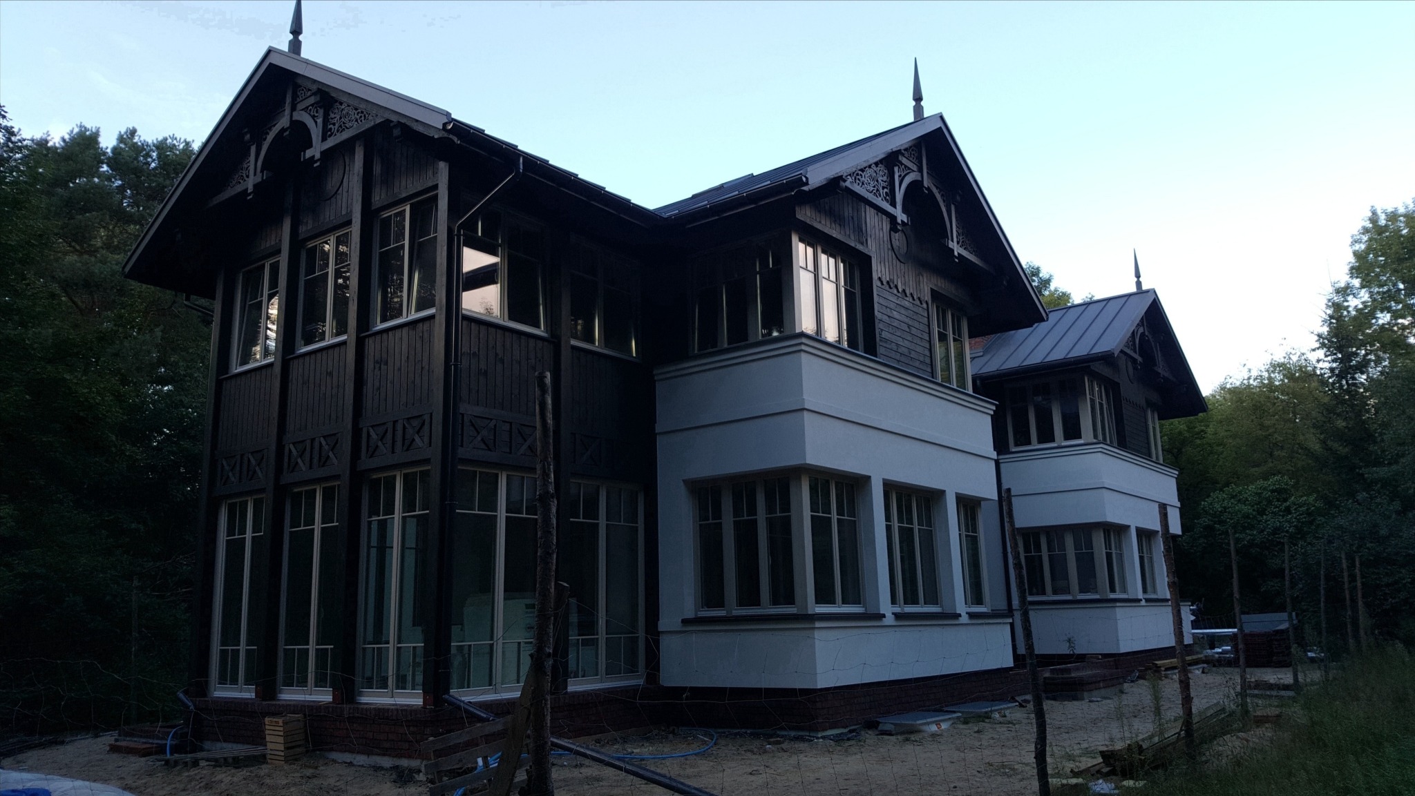 Elewacja dwukondygnacyjnego domu w stylu dworkowym z ciemnego drewna i białego tynku, otoczonego zielenią. Widoczne detale architektoniczne i elementy wykończeniowe.