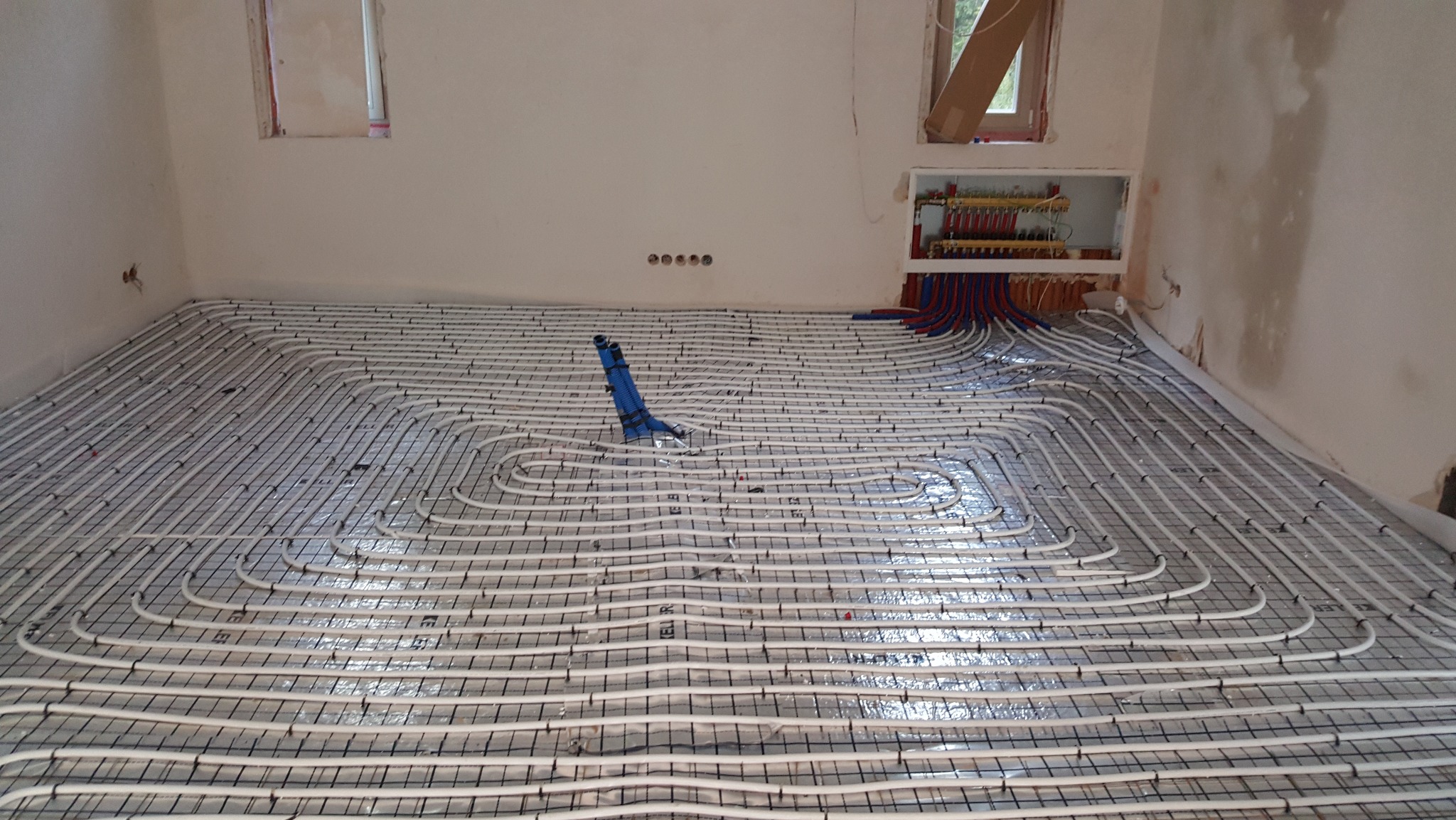 Białe rury ogrzewania podłogowego ułożone na folii aluminiowej, z niebieskim elementem montażowym i rozdzielaczem w ścianie, w surowym wnętrzu.