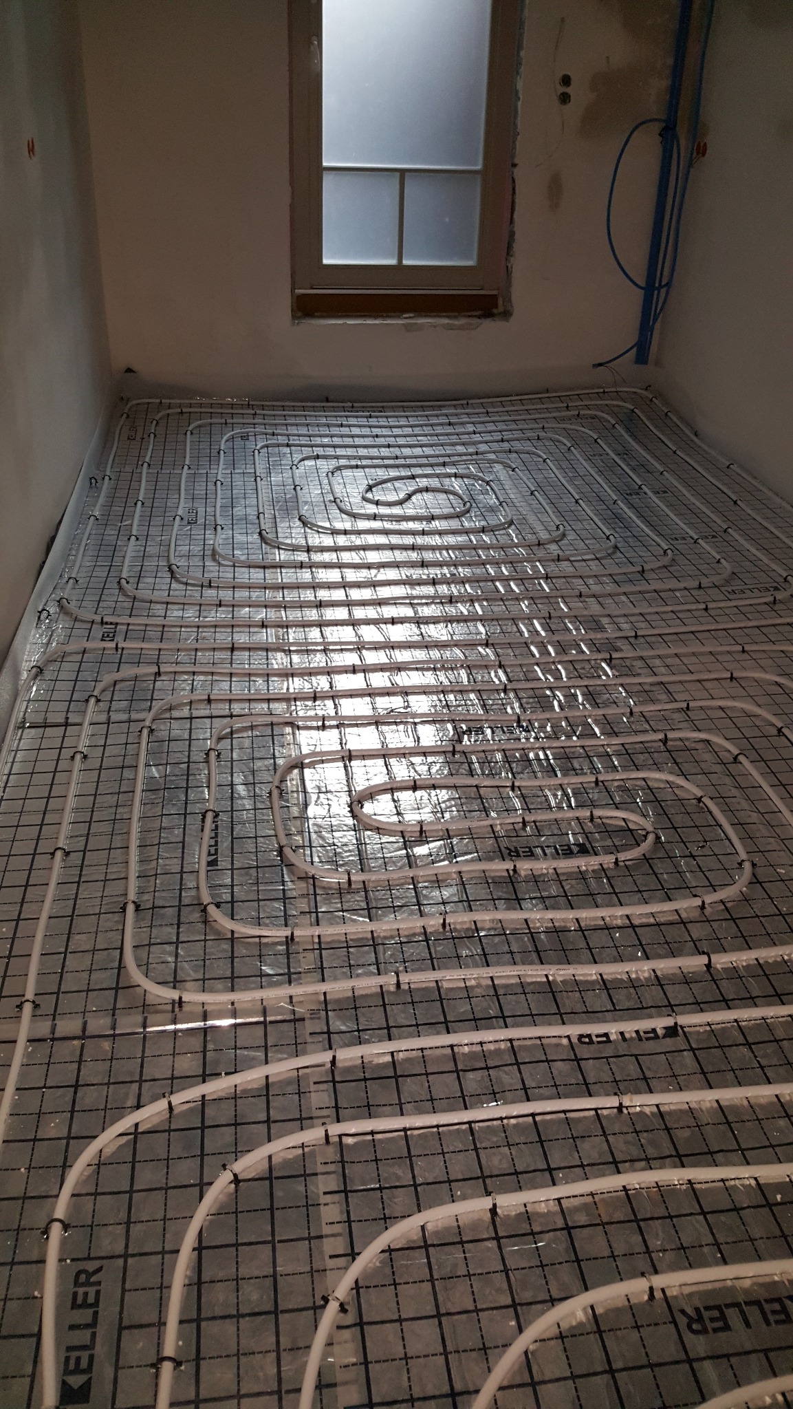 Ułożone rury ogrzewania podłogowego na folii z siatką, widoczne mocowania. W tle okno i fragment instalacji na ścianie. Etap przed wylaniem posadzki.