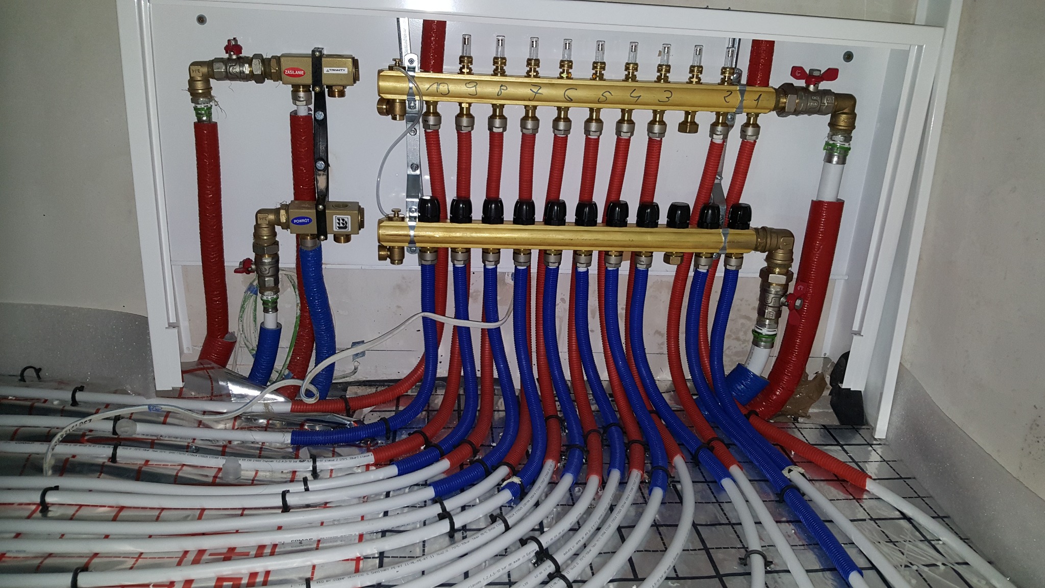 Rozdzielacz ogrzewania podłogowego z rurami w kolorach czerwonym i niebieskim, zamontowany w białej szafce, widoczne białe rury na podłodze z siatką.