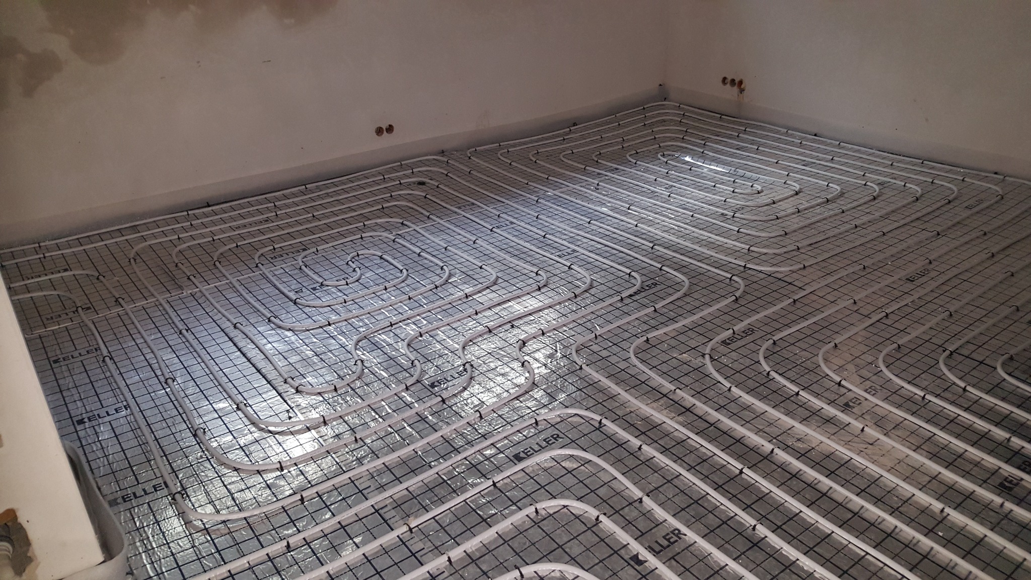 Białe rury ogrzewania podłogowego ułożone na srebrnej folii z nadrukiem siatki i logo Keller, widok z góry, surowy stan ścian.