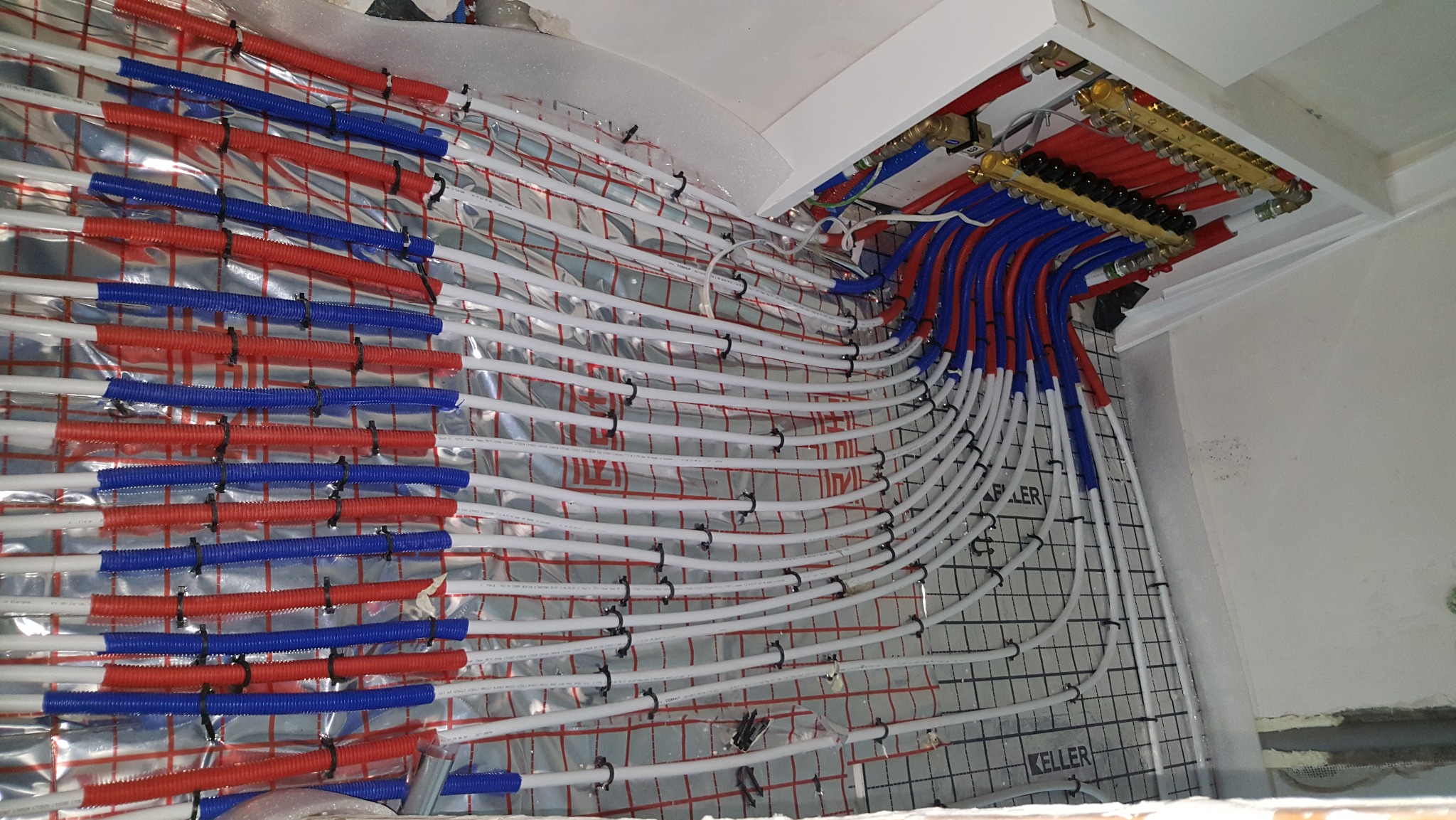 Instalacja ogrzewania podłogowego: rury w izolacji, rozdzielacz z zaworami, widok z góry. Czerwone i niebieskie rury prowadzone równolegle, przymocowane do maty.