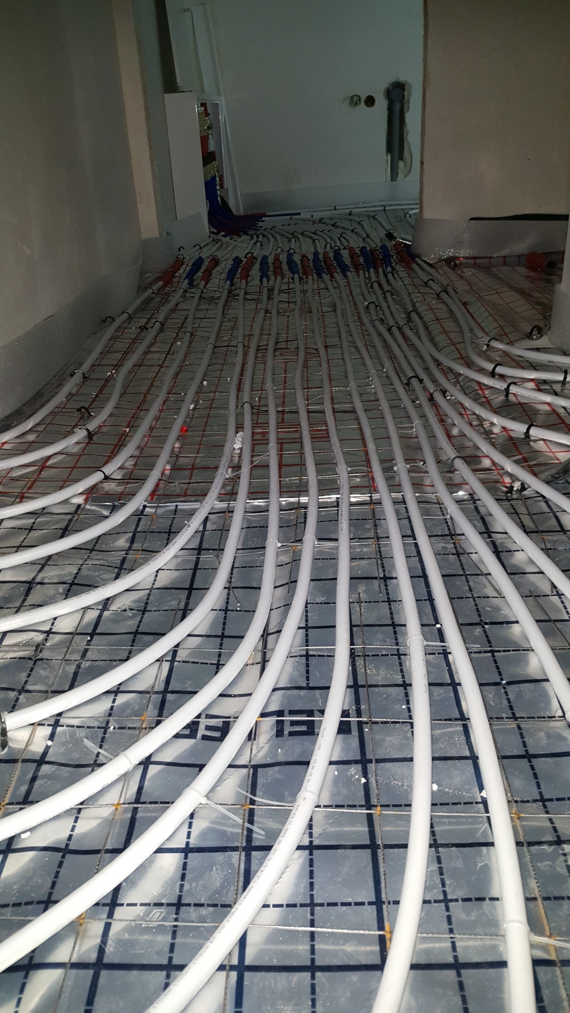 Białe rury ogrzewania podłogowego ułożone na siatce zbrojeniowej, widoczne złącza i fragment rozdzielacza w tle. Instalacja przed wylaniem posadzki.