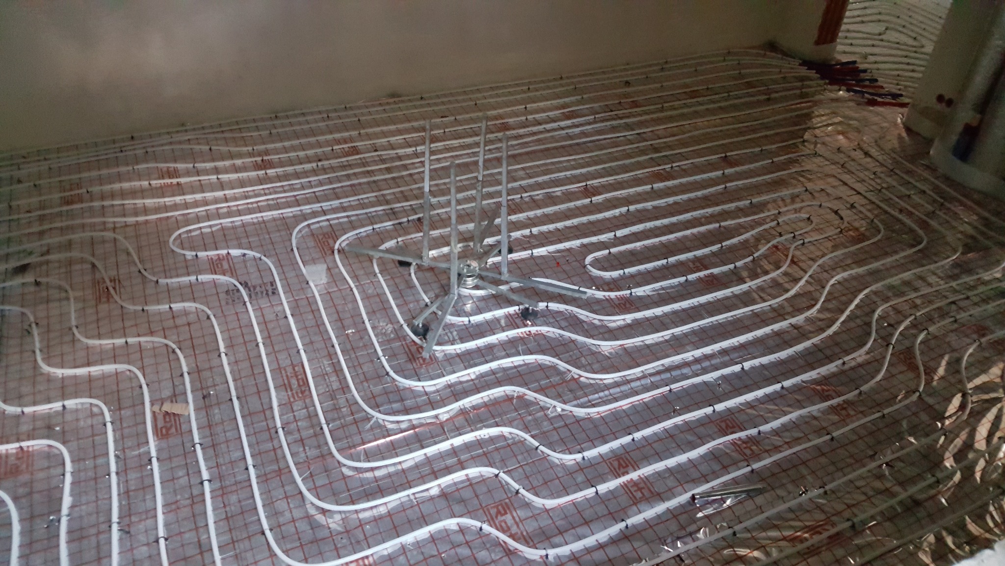 Białe rurki ogrzewania podłogowego ułożone na folii z czerwoną siatką, widoczne mocowania i rozdzielacz. Etap instalacji przed wylaniem posadzki.