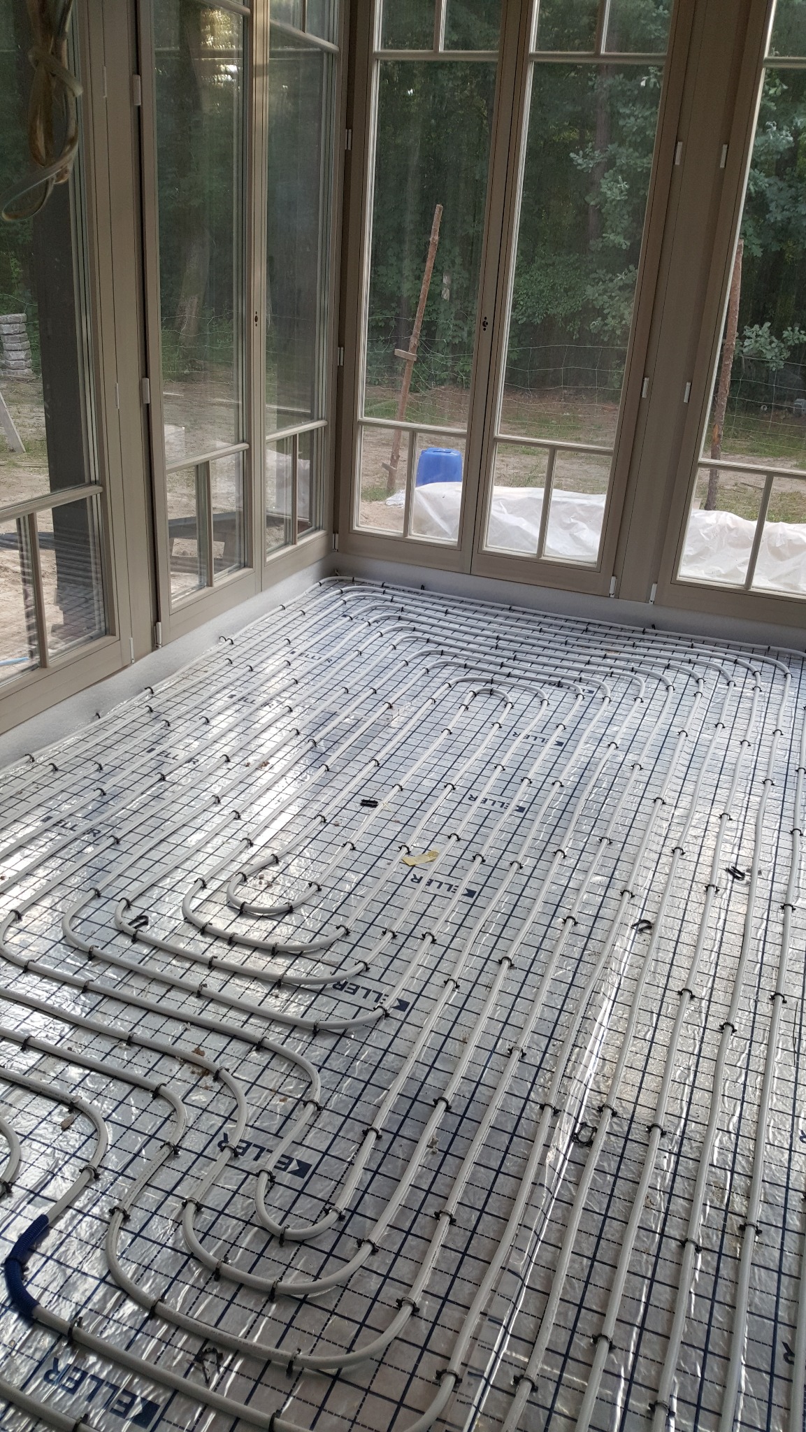 Białe rury ogrzewania podłogowego ułożone na srebrnej folii w jasnym pomieszczeniu z dużymi oknami, widok z góry, etap instalacji przed wylewką.