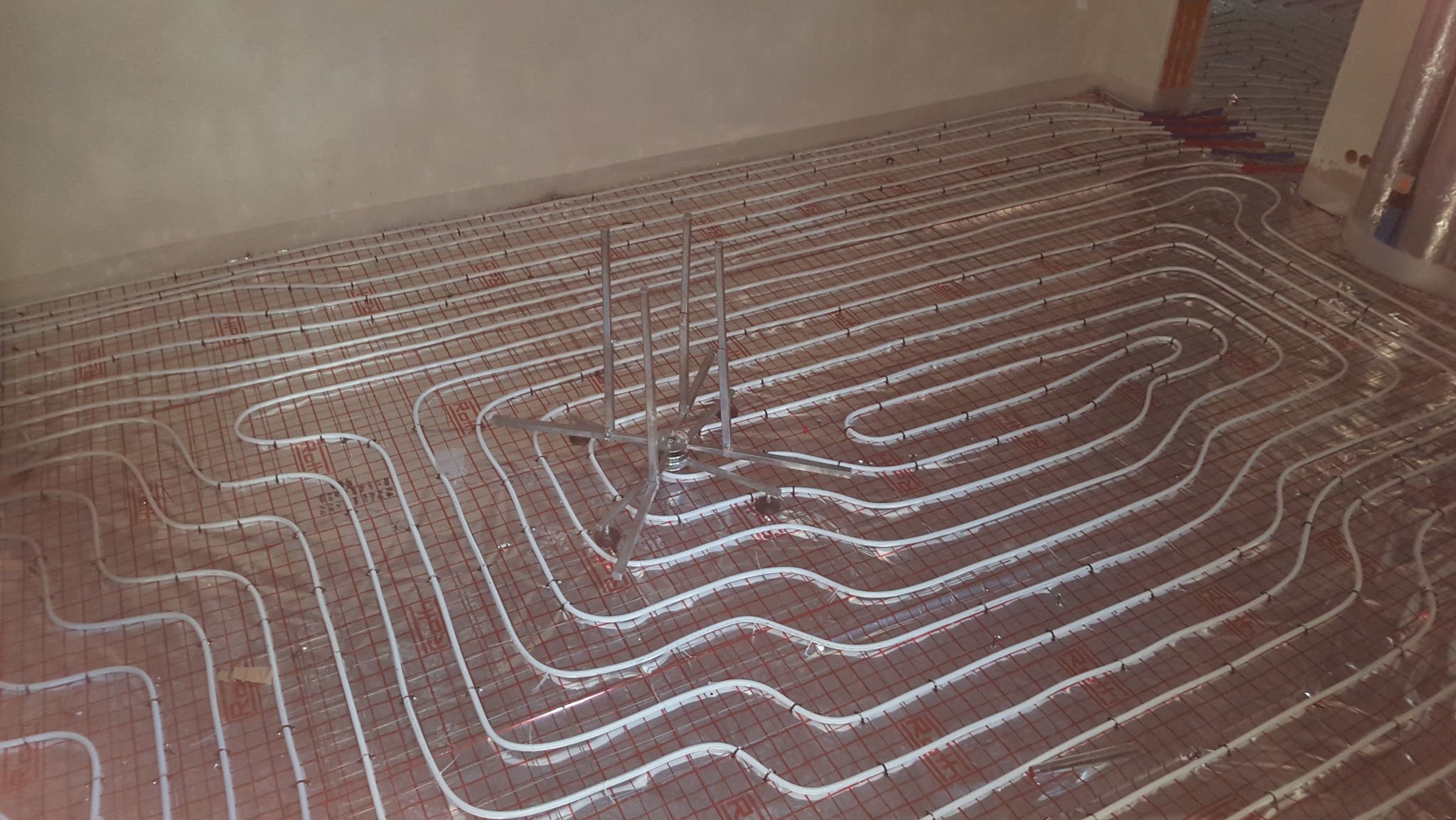 Białe rury ogrzewania podłogowego ułożone na czerwonej siatce, widoczne metalowe uchwyty mocujące, przygotowane do zalania posadzką.