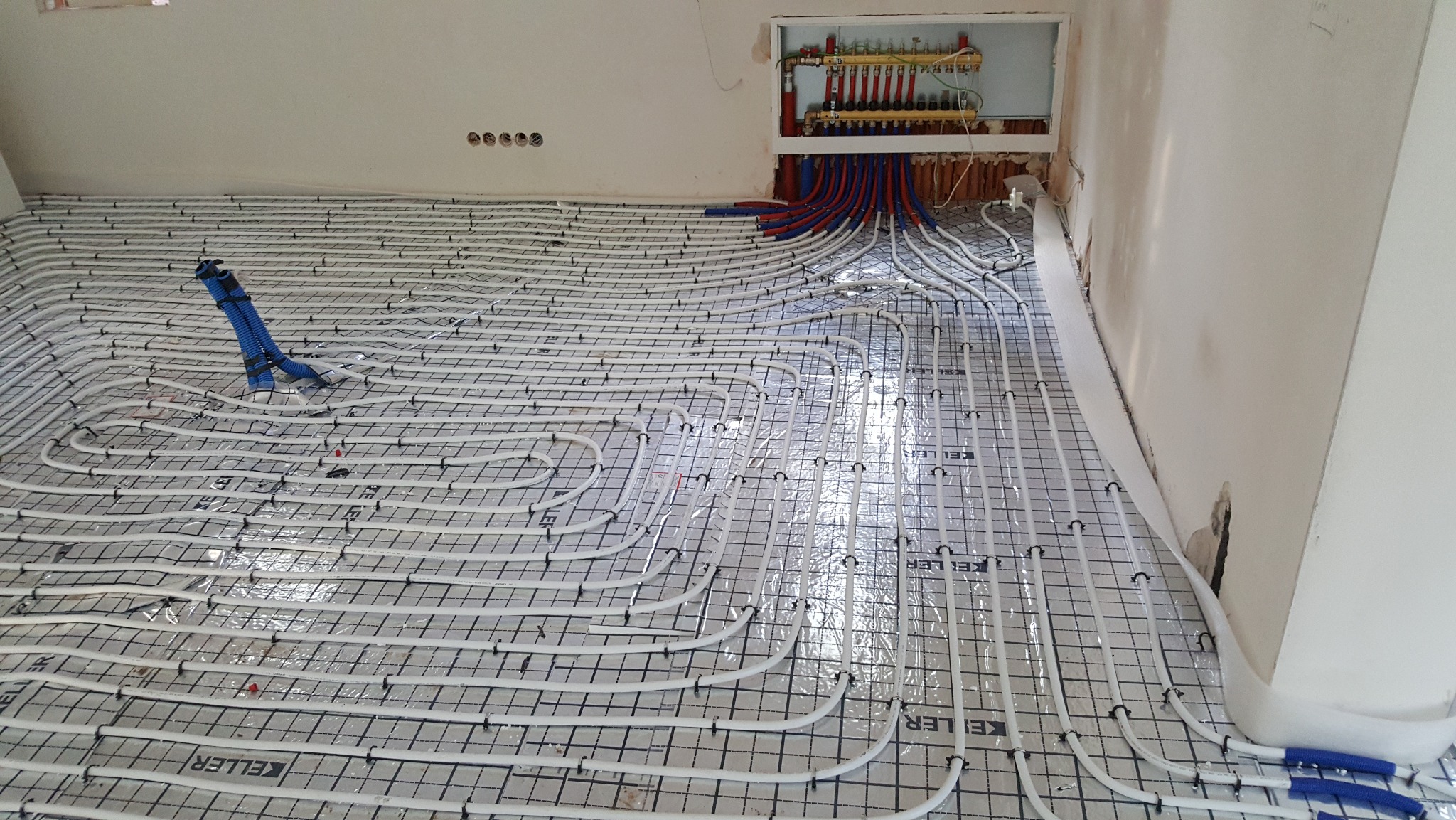 Białe rury ogrzewania podłogowego ułożone na folii z rastrem, z widocznym rozdzielaczem w ścianie. Niebieskie narzędzie do montażu rur na podłodze.