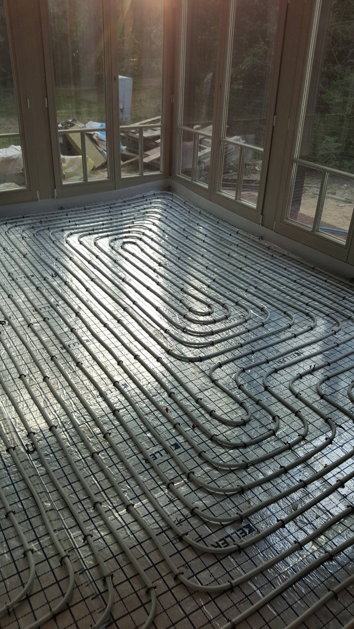 Białe rury ogrzewania podłogowego ułożone spiralnie na folii z siatką w jasnym pomieszczeniu z dużymi oknami, widoczne promienie słoneczne.