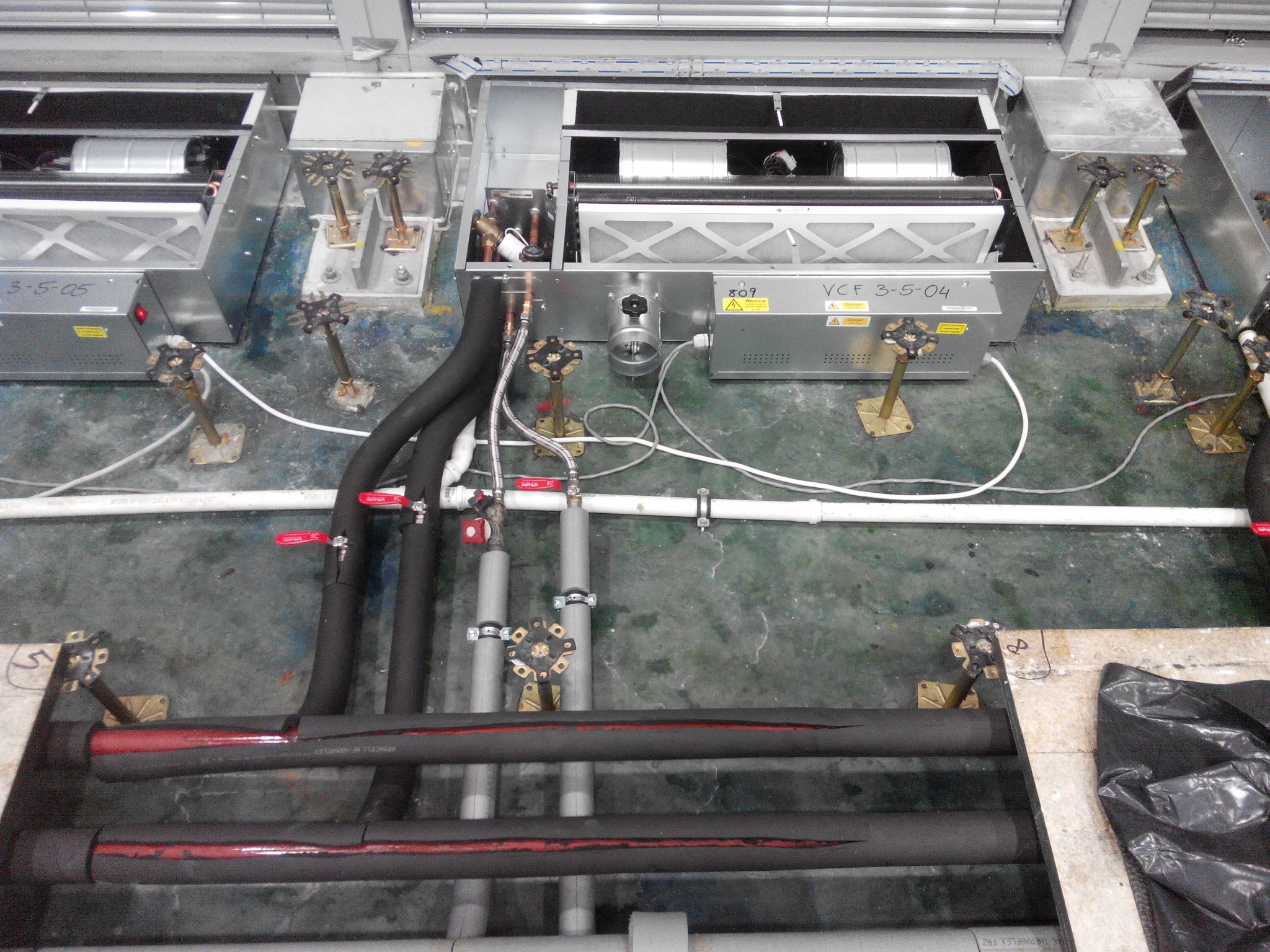 Rozbudowana instalacja hydrauliczna z rurami w izolacji, zaworami i metalowymi elementami mocującymi na szorstkiej, betonowej podłodze. Widoczne numery seryjne na urządzeniach.