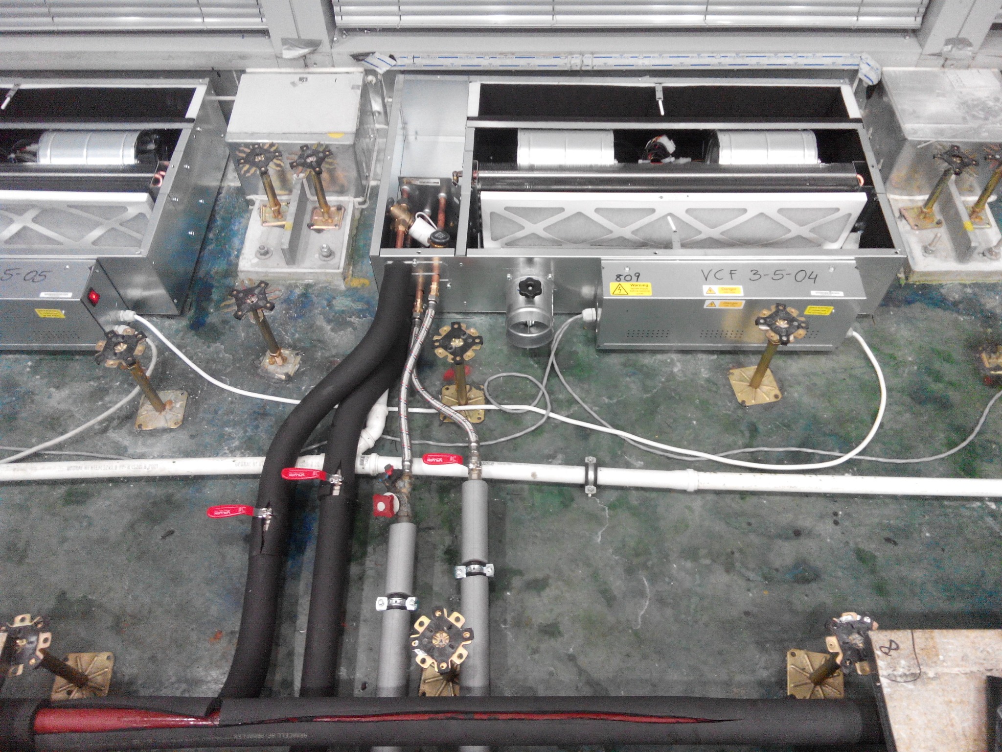 Instalacja przemysłowa z rurami, zaworami i metalowymi skrzynkami. Widoczne elementy wentylacji i klimatyzacji. Szara podłoga z widocznymi śladami użytkowania.