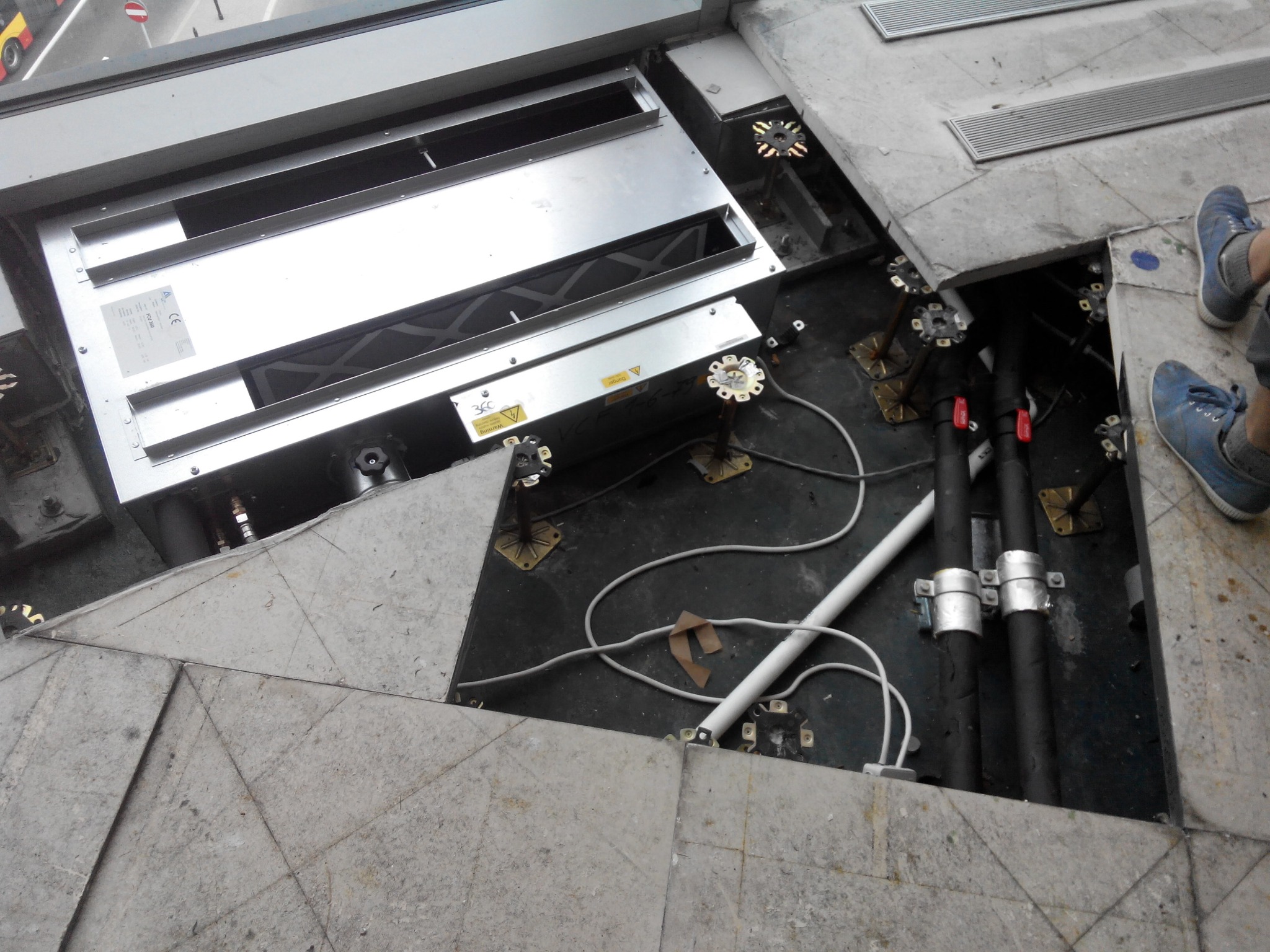 Otwarta podłoga techniczna z widoczną instalacją: rury, kable i metalowa skrzynka. Fragment buta w rogu kadru. Widok z góry na instalację pod podłogą.