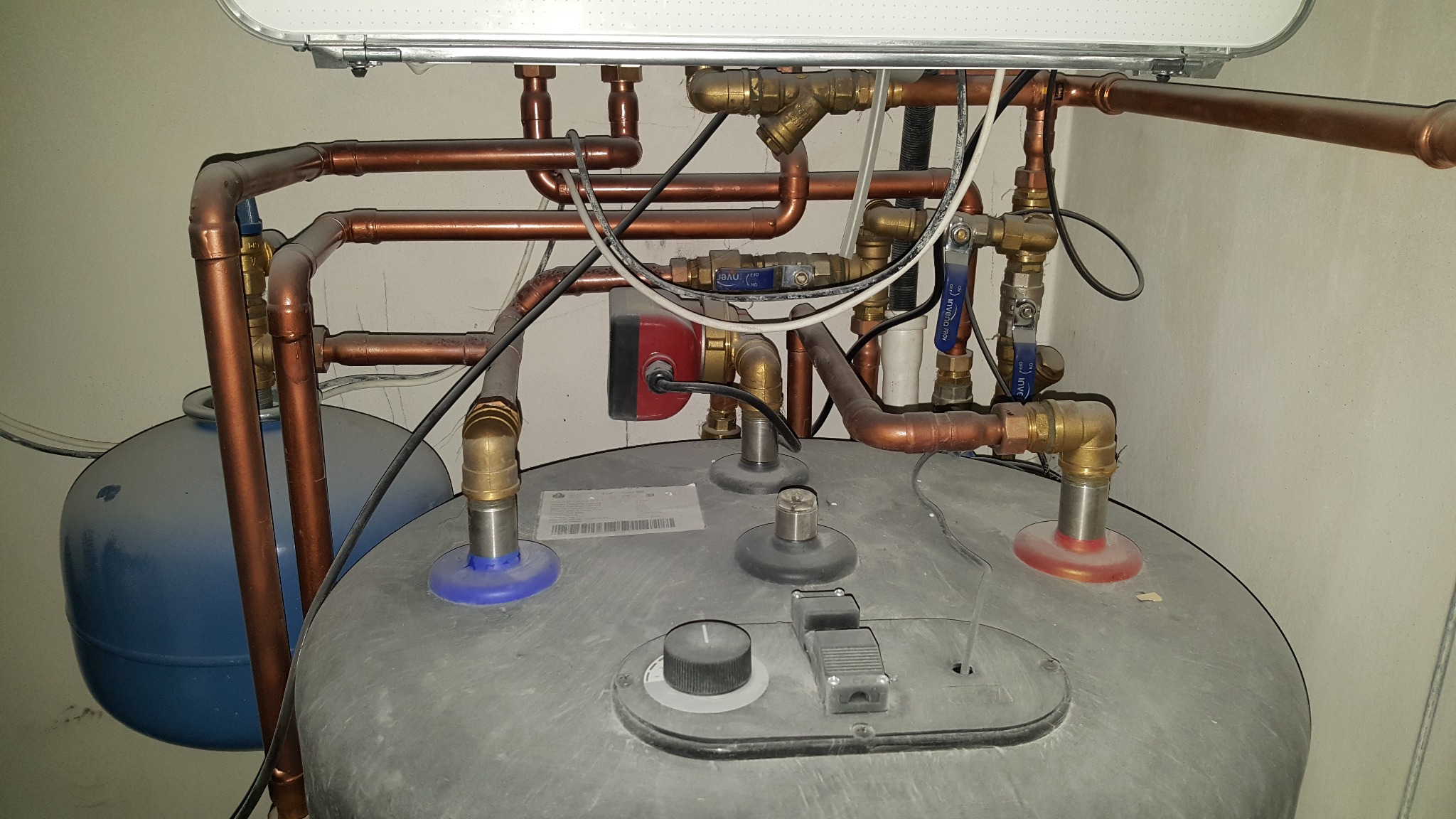 Złożona instalacja grzewcza z miedzianymi rurami, zaworami i pompą obiegową, zamontowana nad szarym zbiornikiem. Widoczne elementy sterujące i niebieski zbiornik wyrównawczy.