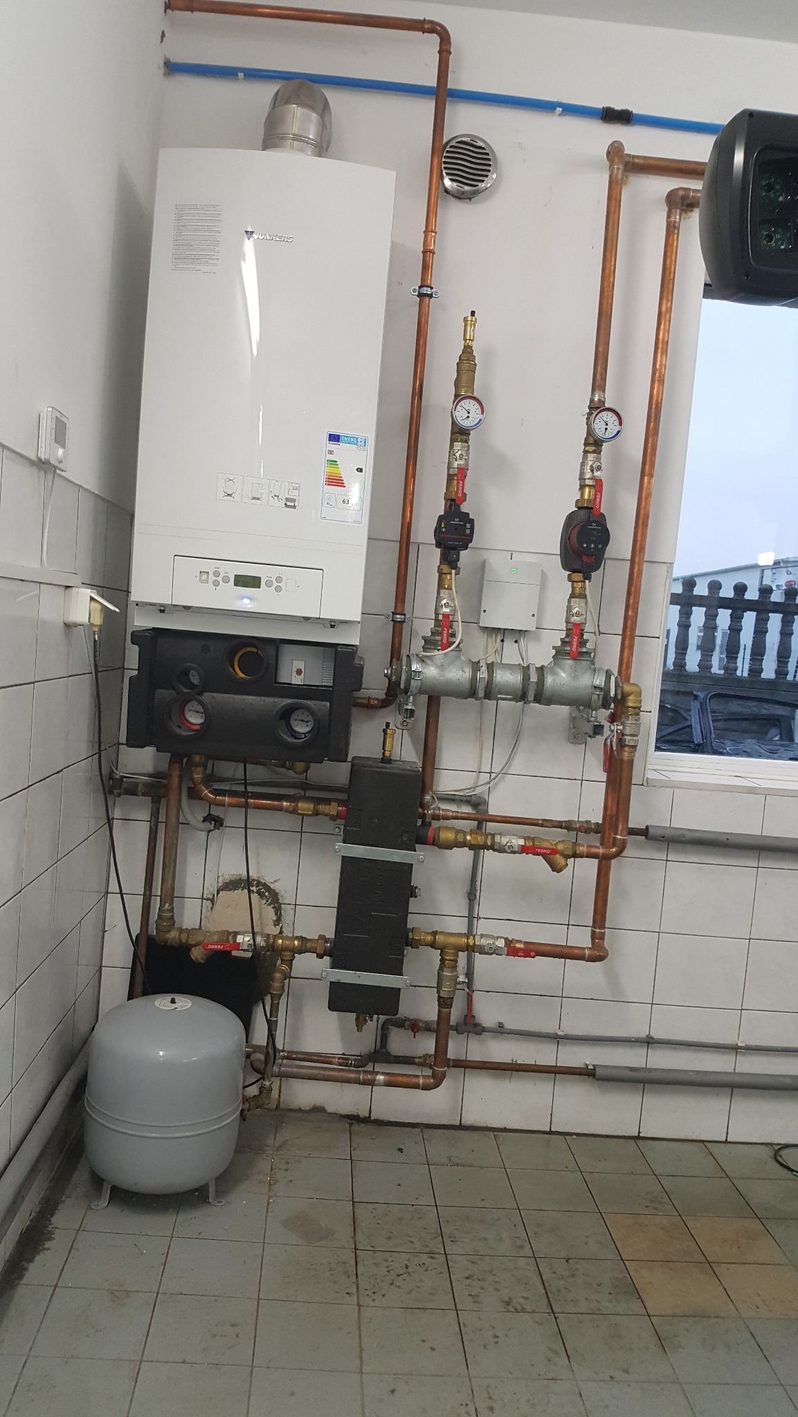 Biały piec gazowy Junkers z miedzianą instalacją, manometrami i szarym zbiornikiem wyrównawczym na tle białych płytek. Widoczne zawory i elementy armatury.
