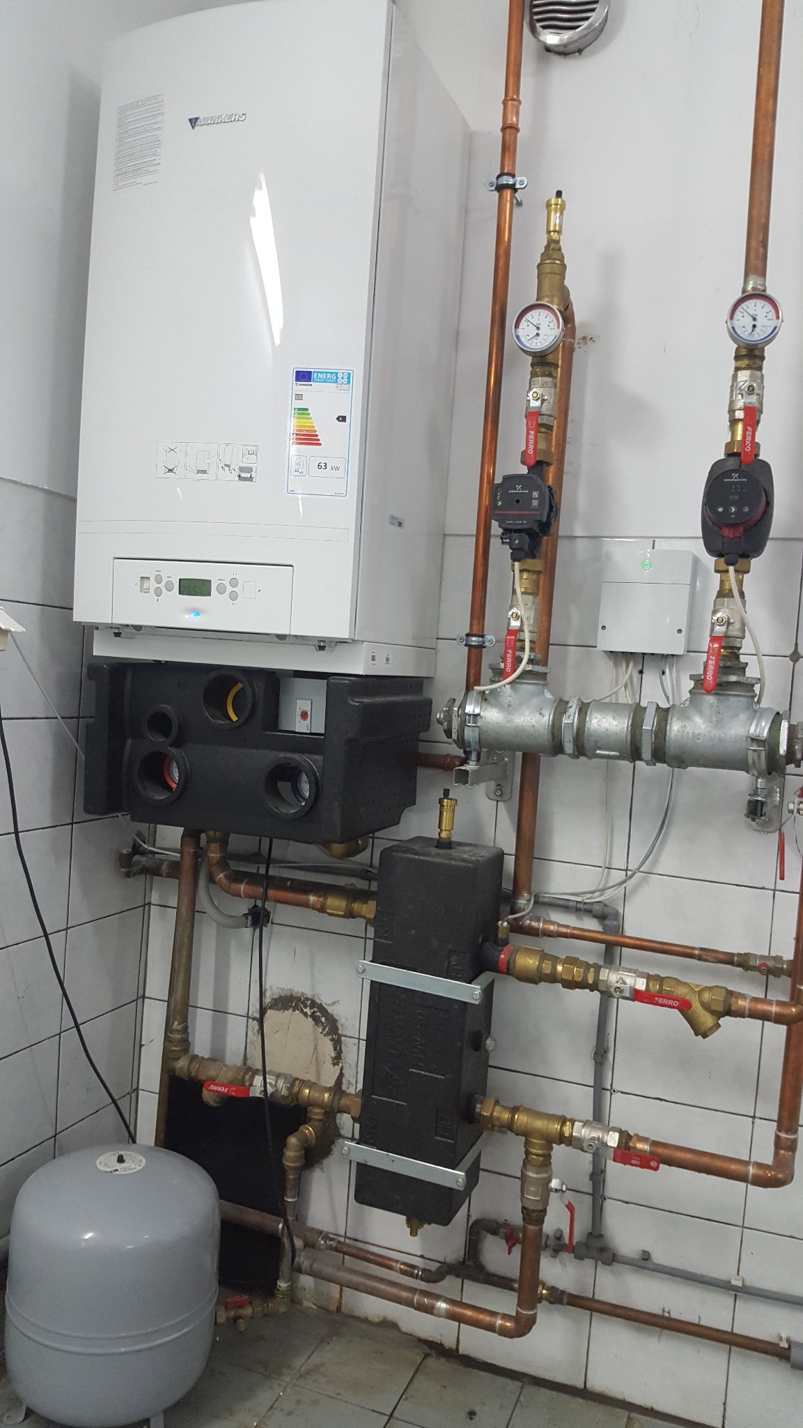 Biały piec gazowy Junkers zamontowany na ścianie z instalacją hydrauliczną z miedzianych rur, zaworami i manometrami. Widoczny szary zbiornik wyrównawczy.