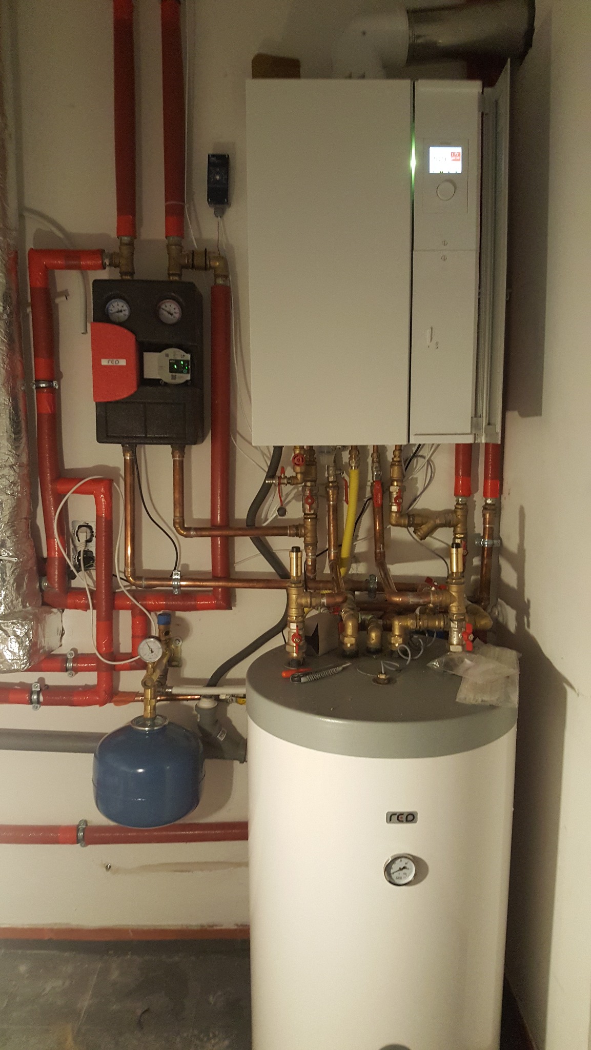 Zmontowany, nowoczesny piec gazowy z zasobnikiem wody, widoczne rury instalacji w kolorze czerwonym i miedzianym, manometry i sterownik.