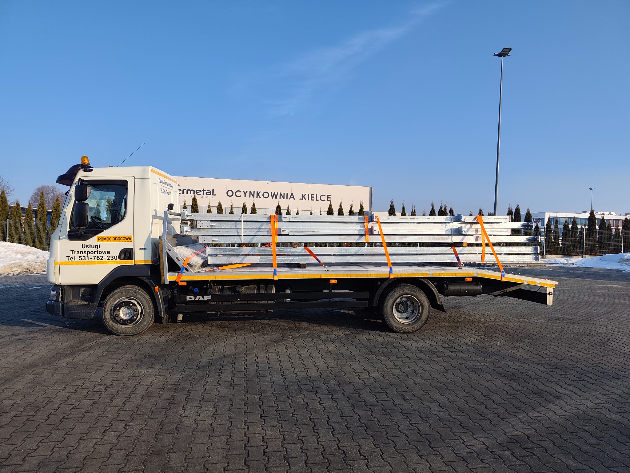 Laweta DAF z ładunkiem profili stalowych zabezpieczonych pasami transportowymi, na tle budynku z napisem 'Ocynkownia Kielce'.