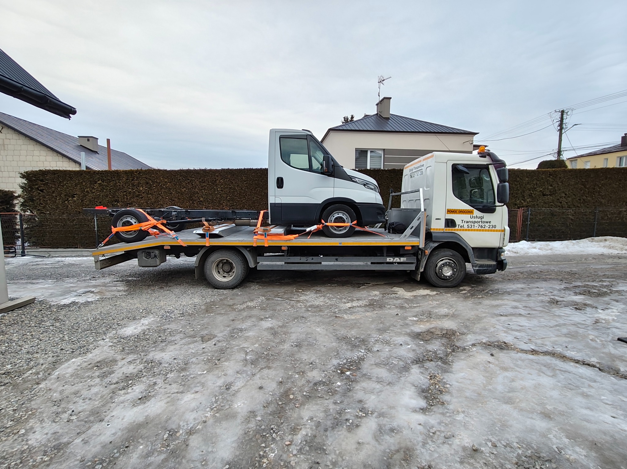 Laweta DAF z białym busem na platformie, zabezpieczonym pomarańczowymi pasami. Widok z boku na tle zabudowań i pochmurnego nieba.