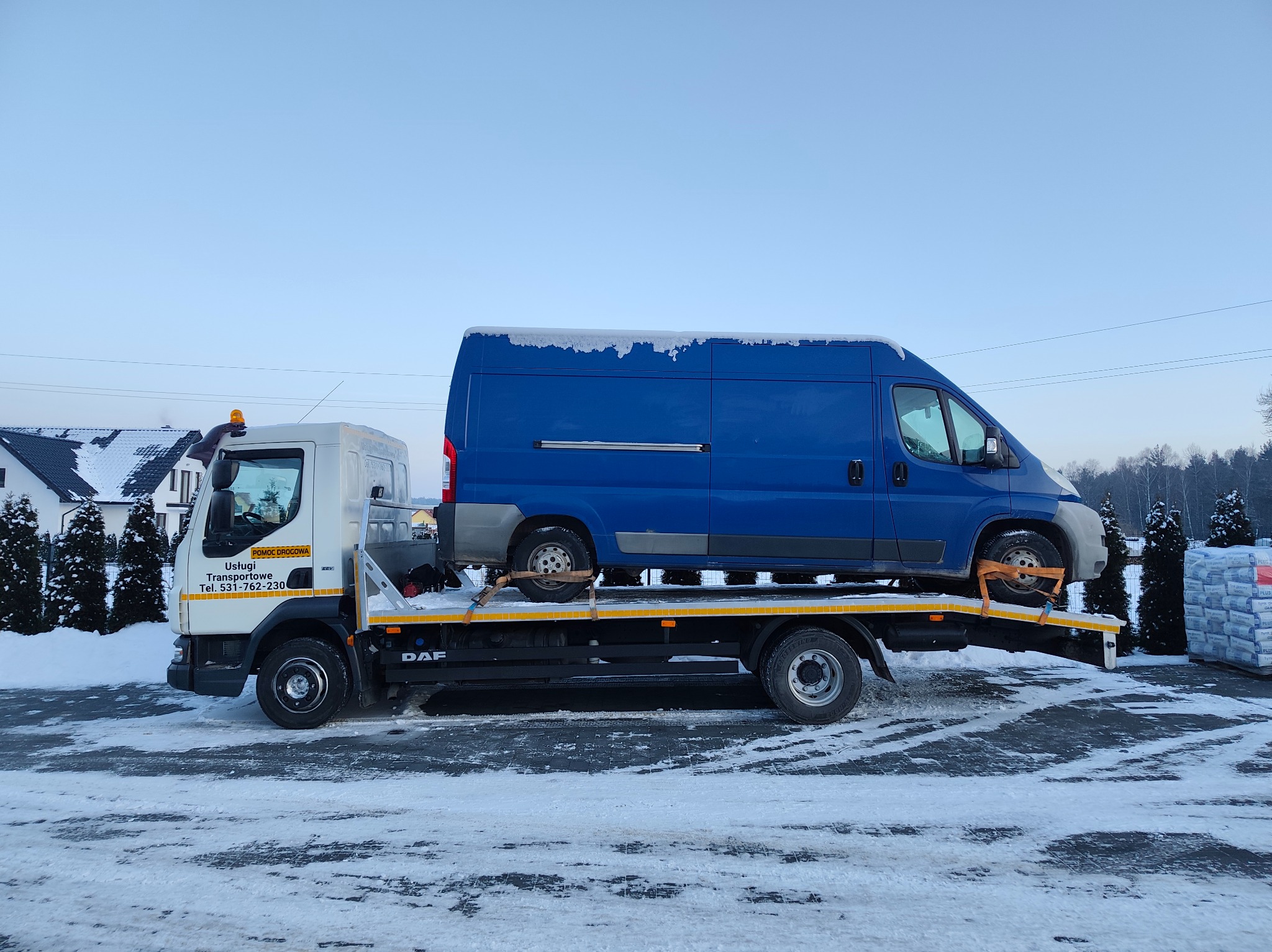 Laweta DAF z niebieskim busem na platformie, zimowy krajobraz. Widoczny budynek w tle i śnieg na dachu busa. Pomoc drogowa z numerem telefonu.