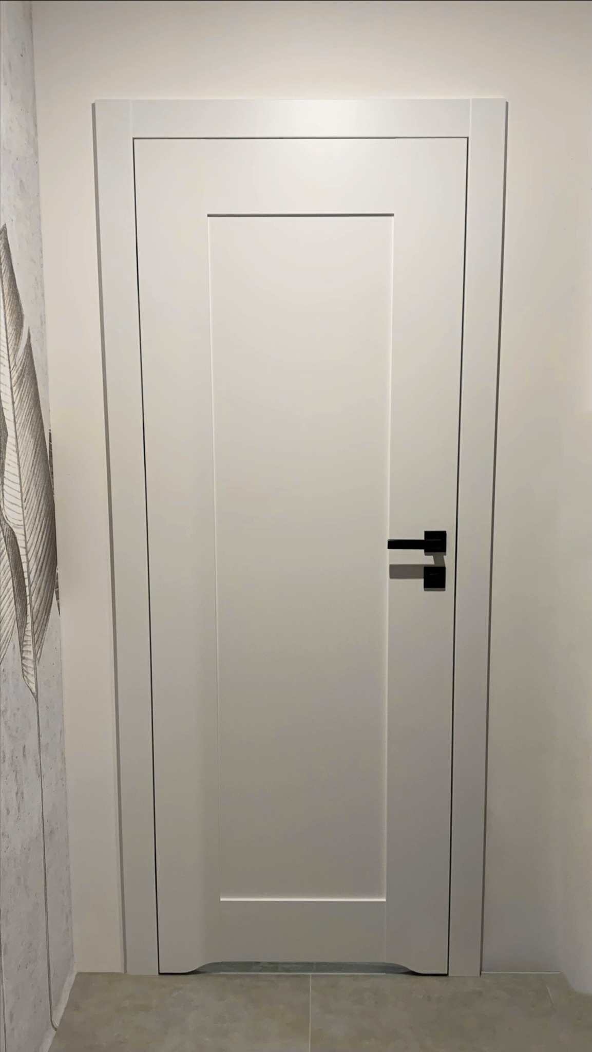 Zamknięte, białe drzwi wewnętrzne z czarną klamką, wkomponowane w jasną ścianę z subtelnym wzorem. Minimalistyczny design, widoczne detale wykończenia.