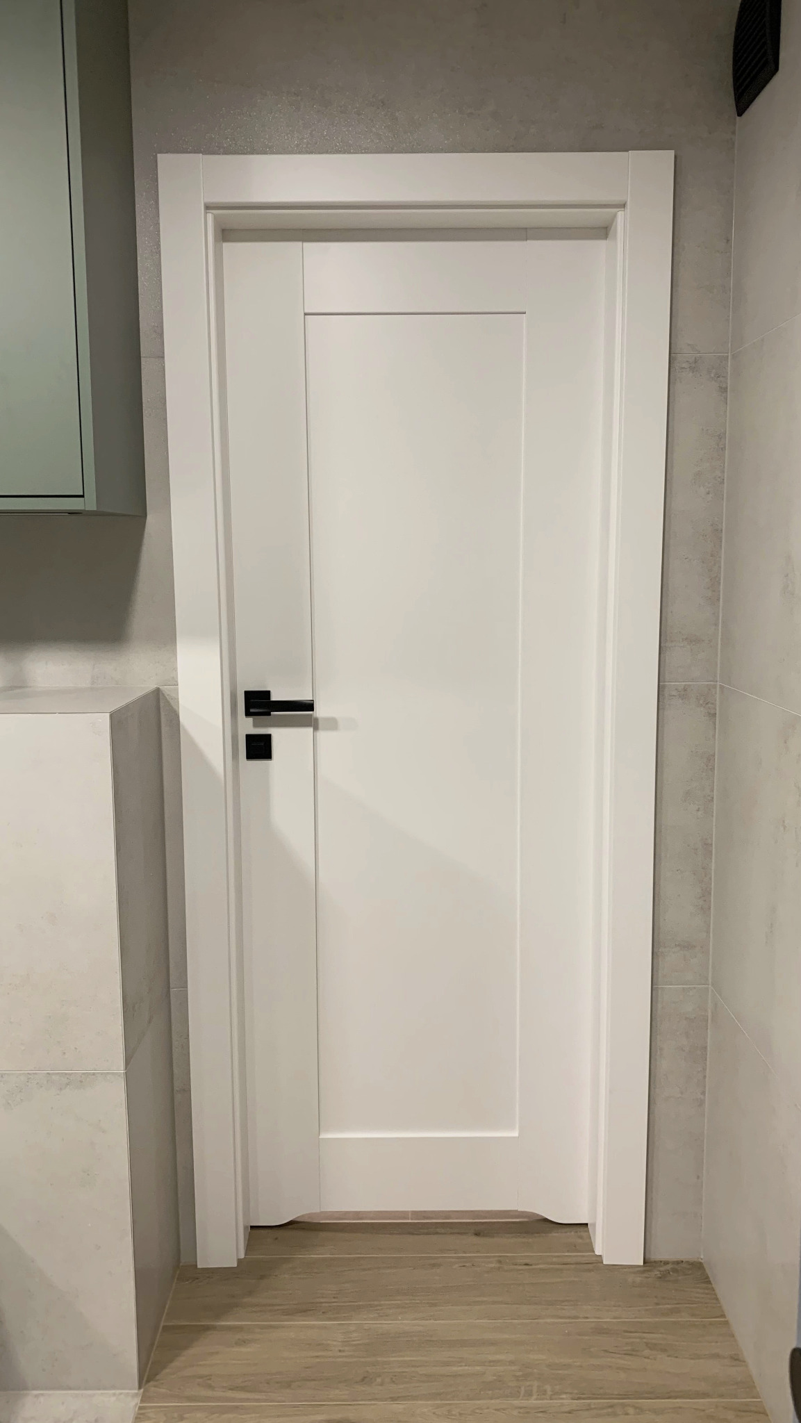 Białe drzwi wewnętrzne z czarną klamką w nowoczesnym wnętrzu, widoczne płytki imitujące kamień na ścianie i drewnopodobna podłoga.