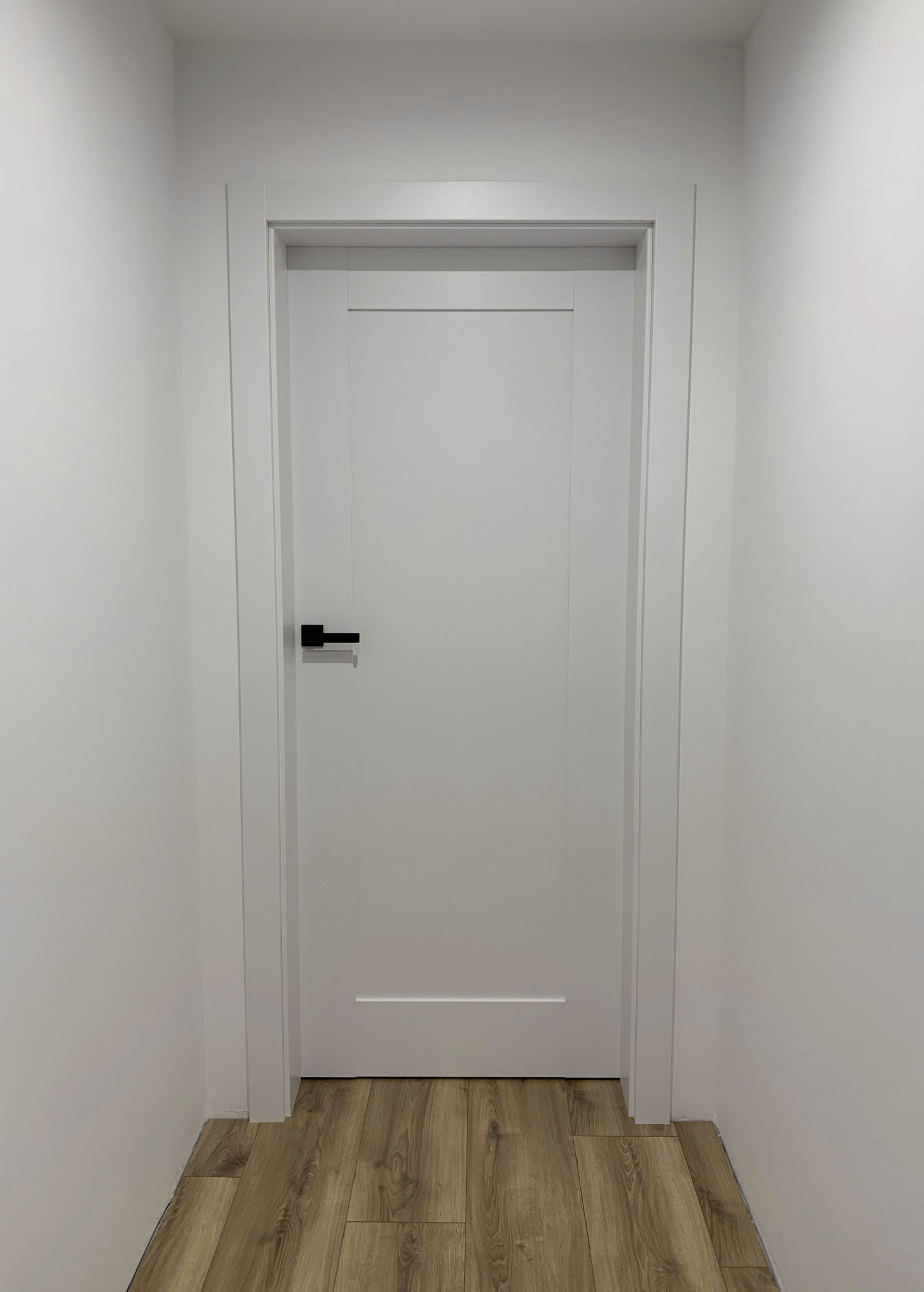 Białe, minimalistyczne drzwi z czarną klamką w białej futrynie, na tle jasnych ścian i podłogi z paneli imitujących drewno. Nowoczesny design.