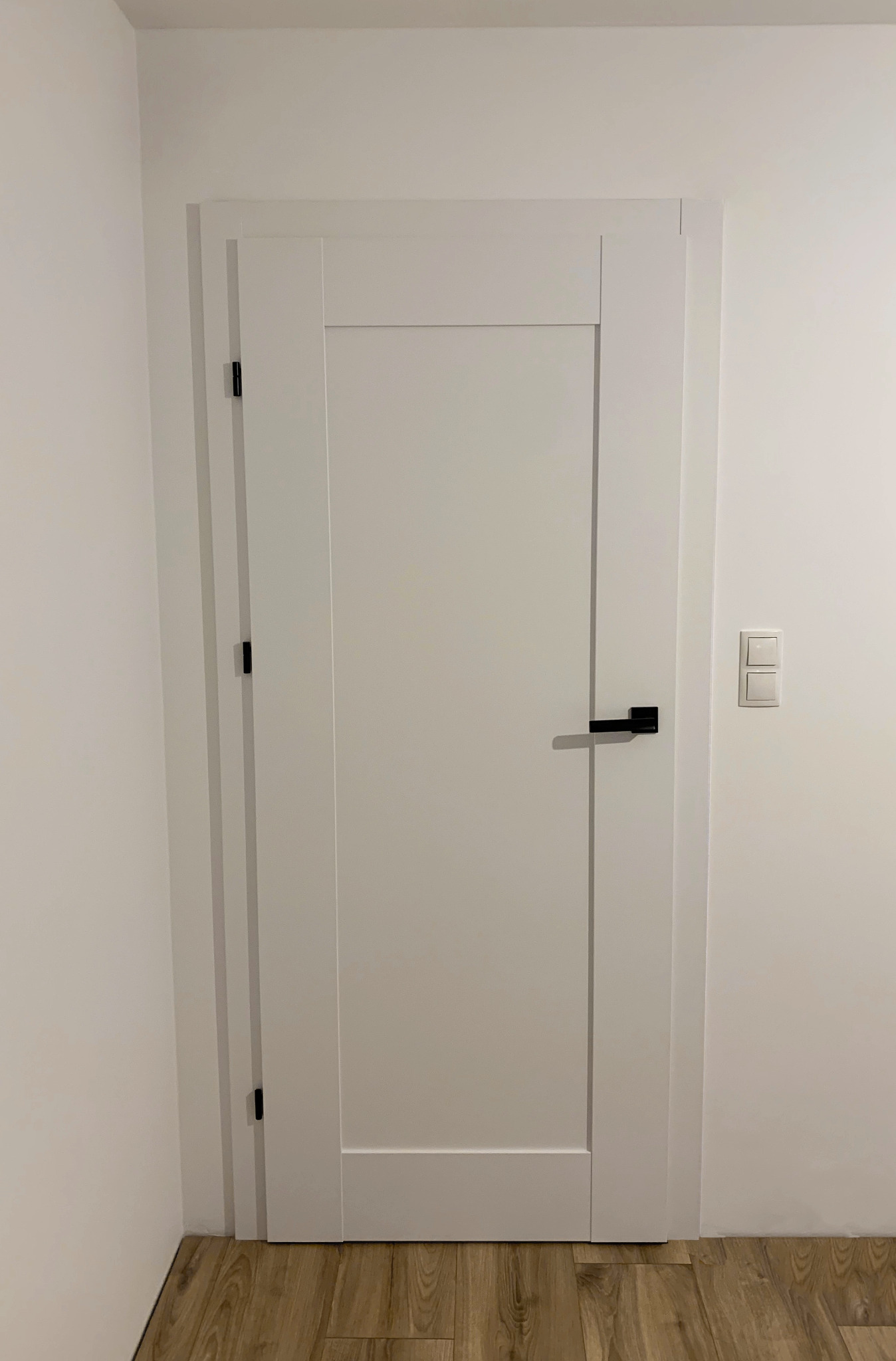 Zamknięte, białe drzwi wewnętrzne z czarną klamką i minimalistyczną ościeżnicą, zamontowane w białej ścianie z włącznikiem światła. Podłoga drewnopodobna.