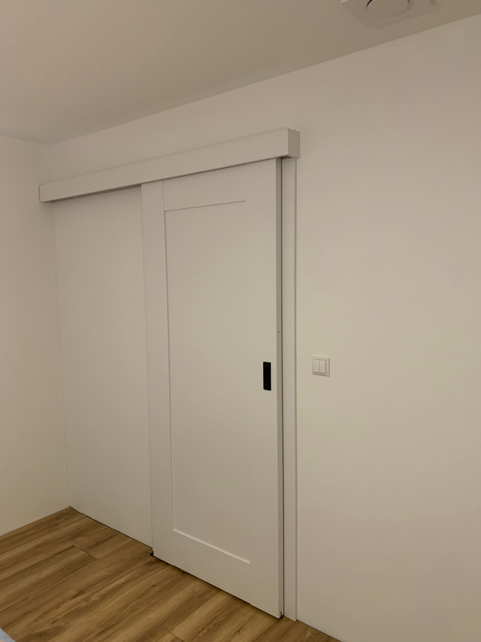Białe, przesuwne drzwi z czarnym uchwytem, zamontowane w jasnym pomieszczeniu z panelami podłogowymi. Widoczny włącznik światła na ścianie.