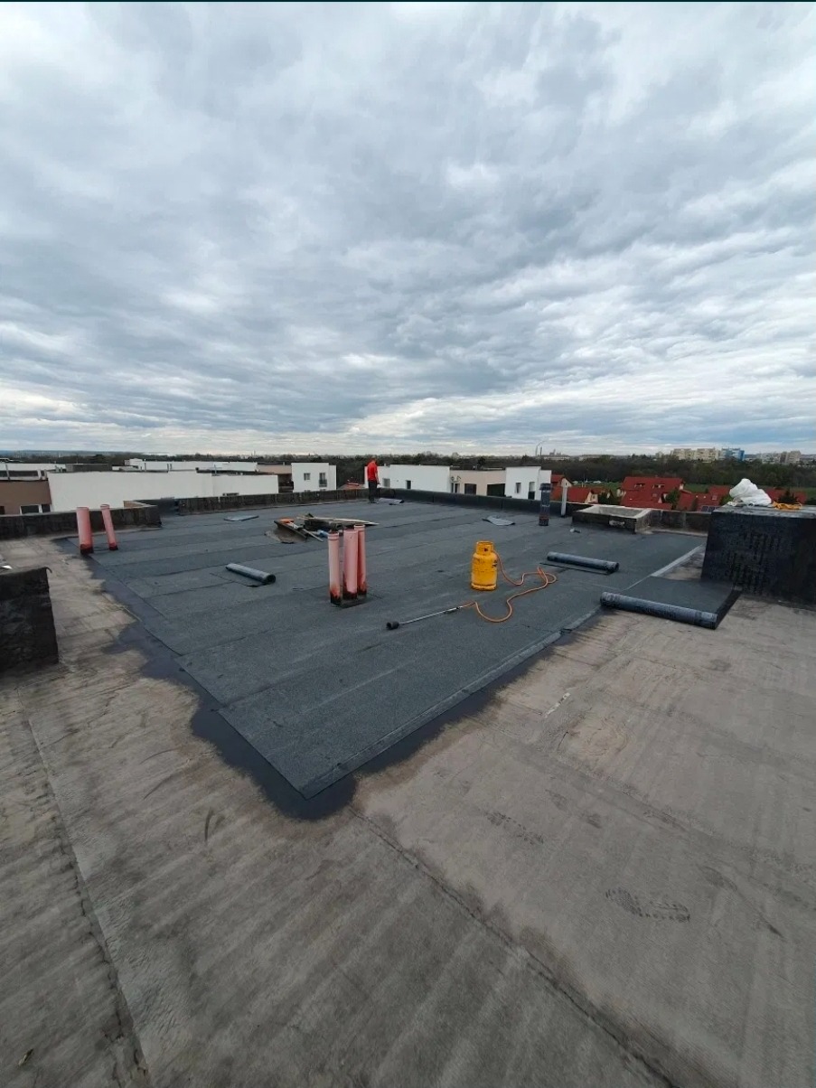 Wykończony dach płaski z papy termozgrzewalnej, widoczne kominy wentylacyjne, butla gazowa i narzędzia dekarskie. W tle zabudowa miejska pod pochmurnym niebem.