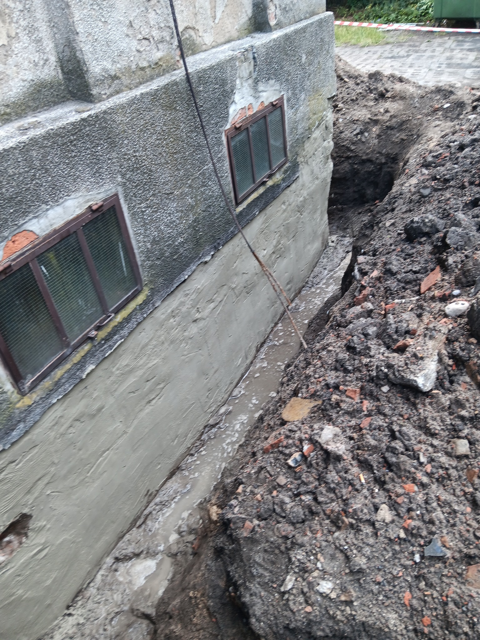 Odkopany fundament budynku z nałożoną warstwą szpachli, widoczne okna piwniczne i wykop z ziemią. Prace ziemne i zabezpieczenie fundamentów.